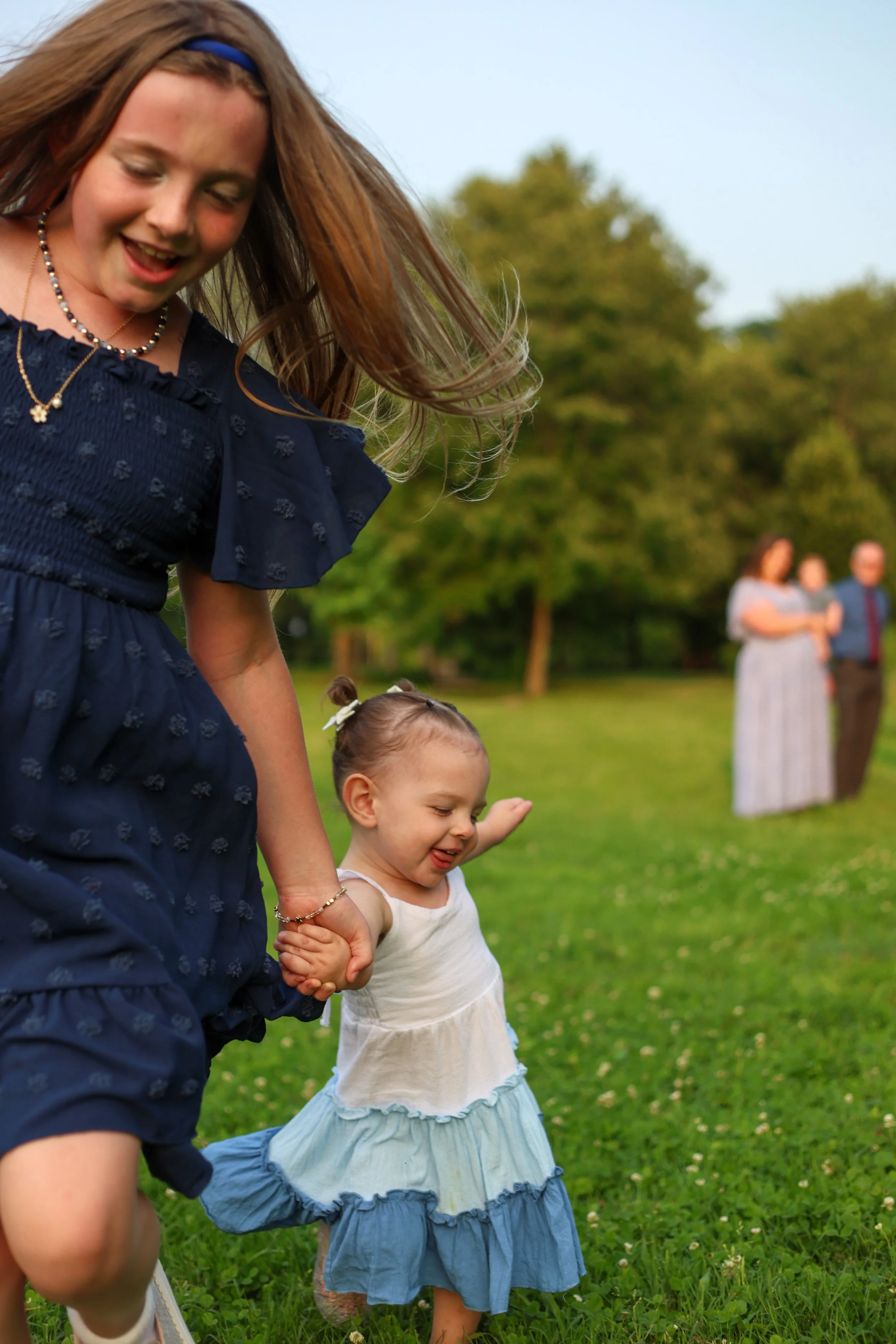 A young girl in a white dress with a blue ruffled skirt holding hands with an older girl in a navy dress, both walking on a grassy field during daytime, with trees and three women talking in the background.