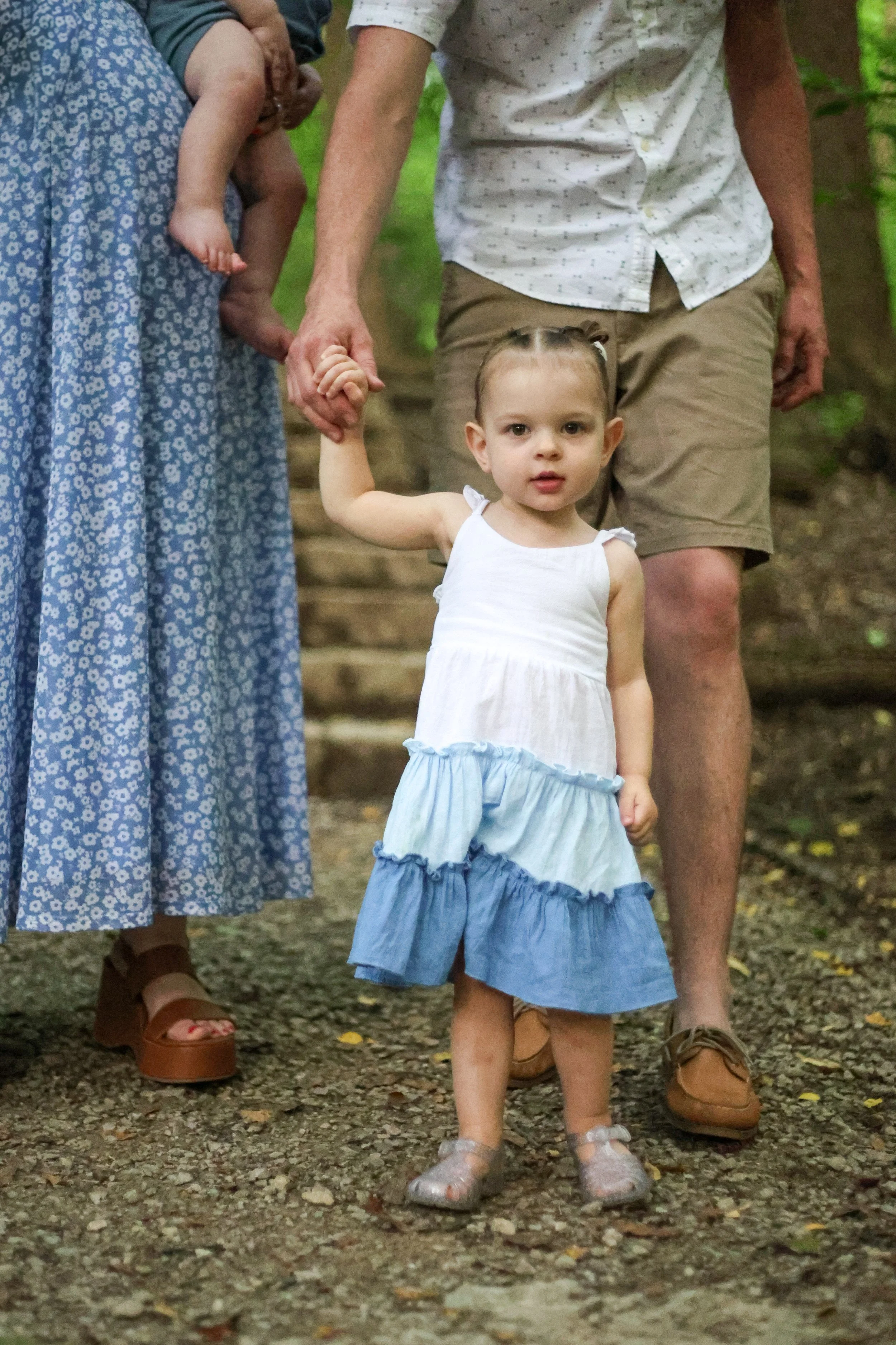 A young girl in a white and blue dress standing outdoors on a dirt path, holding hands with an adult woman and man, surrounded by trees.