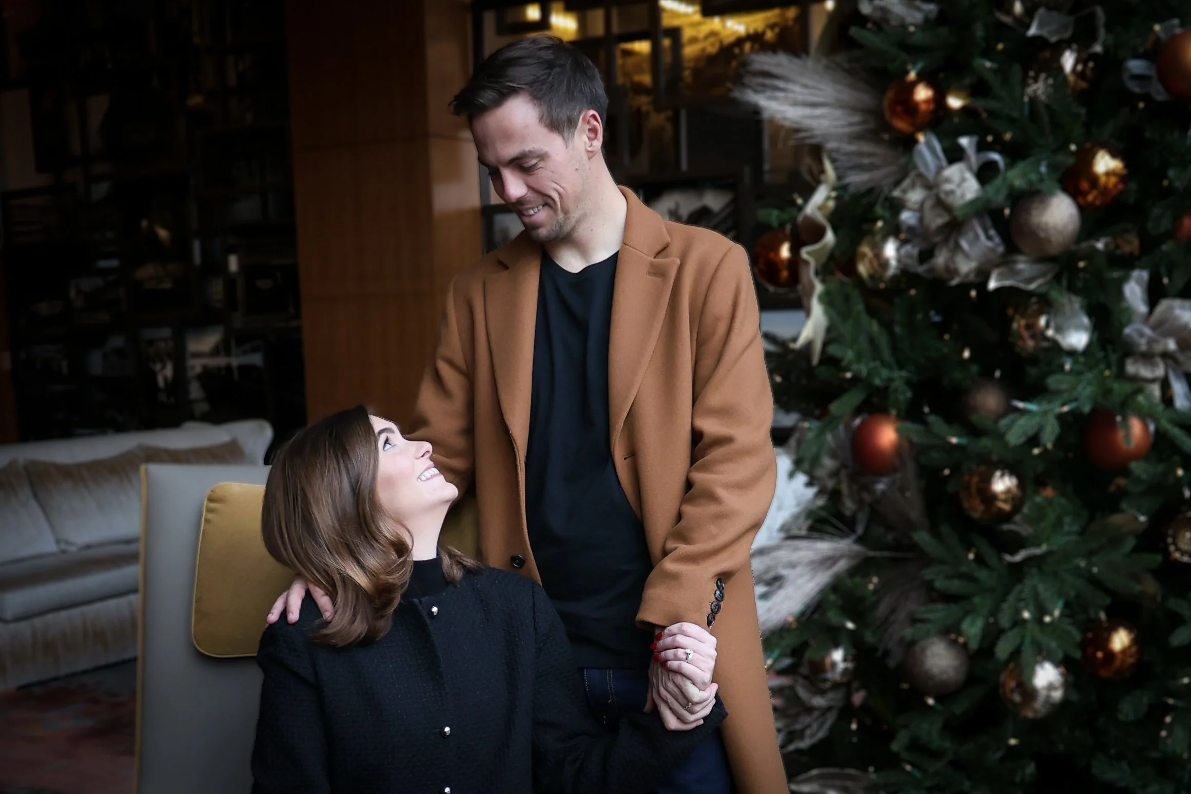 A man in a tan coat and black shirt smiling at a woman with red hair in a black sweater, sitting on a yellow chair, during Christmas decorated with a large decorated Christmas tree in the background.