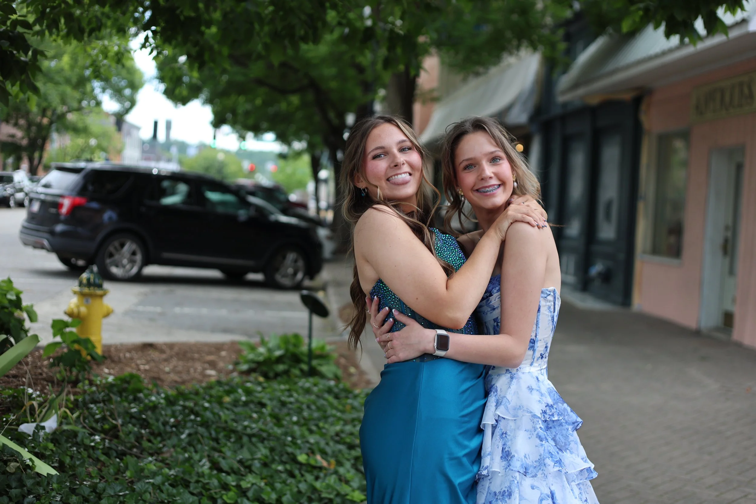 Two young women are standing outdoors on a sidewalk, hugging and smiling at the camera. One woman is wearing a blue dress, and the other is wearing a blue and white floral dress. There are trees, parked cars, and storefronts in the background.