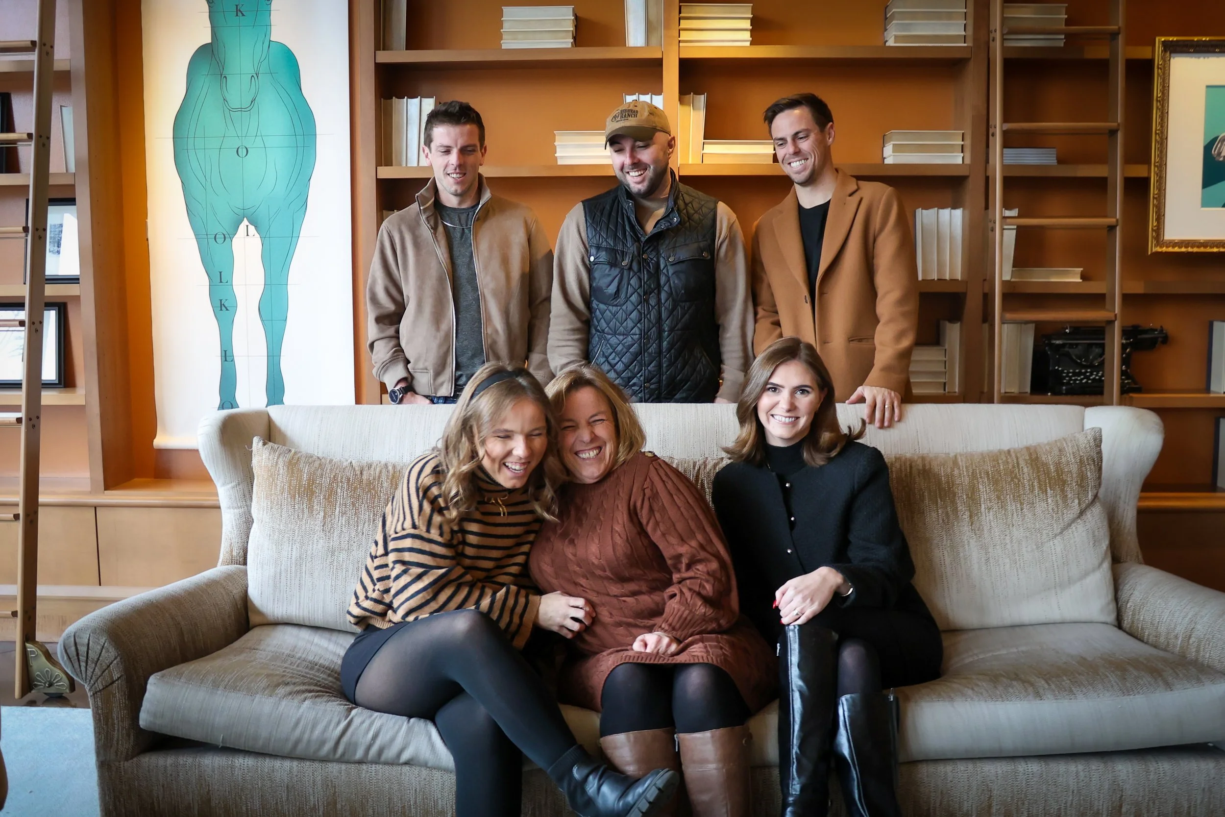 A group of three men standing behind a sofa and three women sitting on it, all smiling and enjoying each other's company in a cozy room with bookshelves and a blue diagram of the human body on the wall.