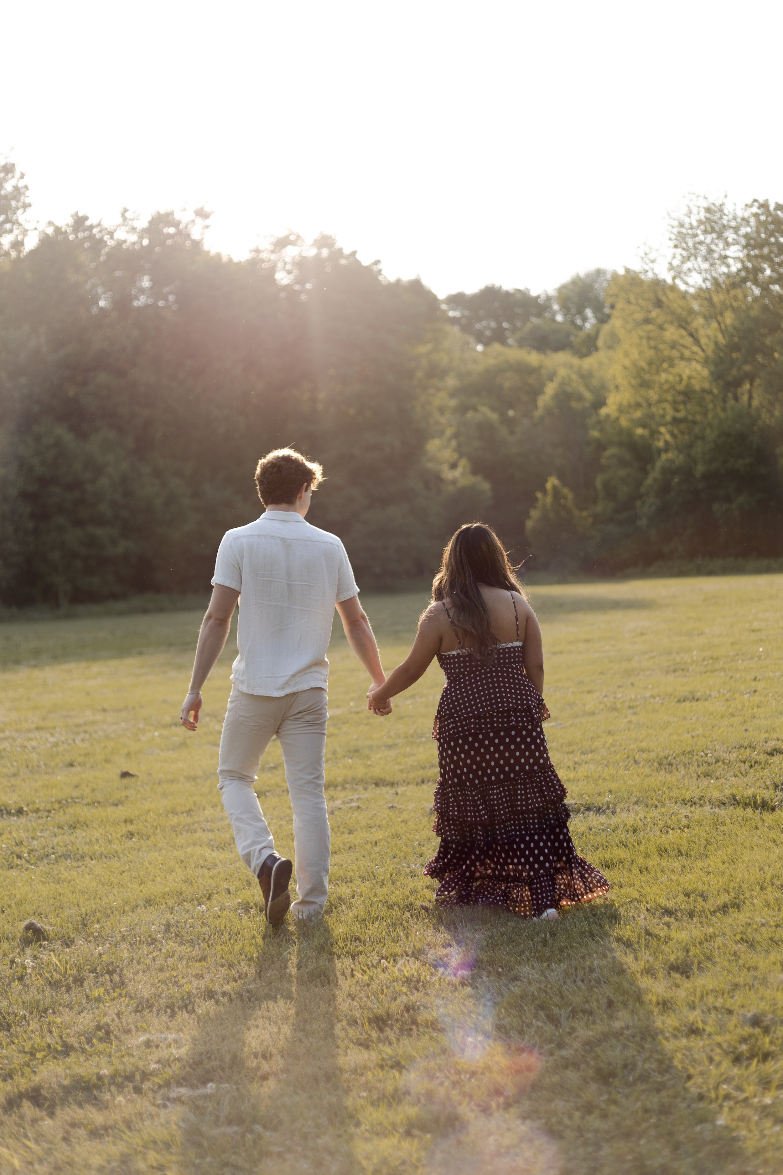 A young couple holding hands and walking through a grassy field with trees in the background during sunset.