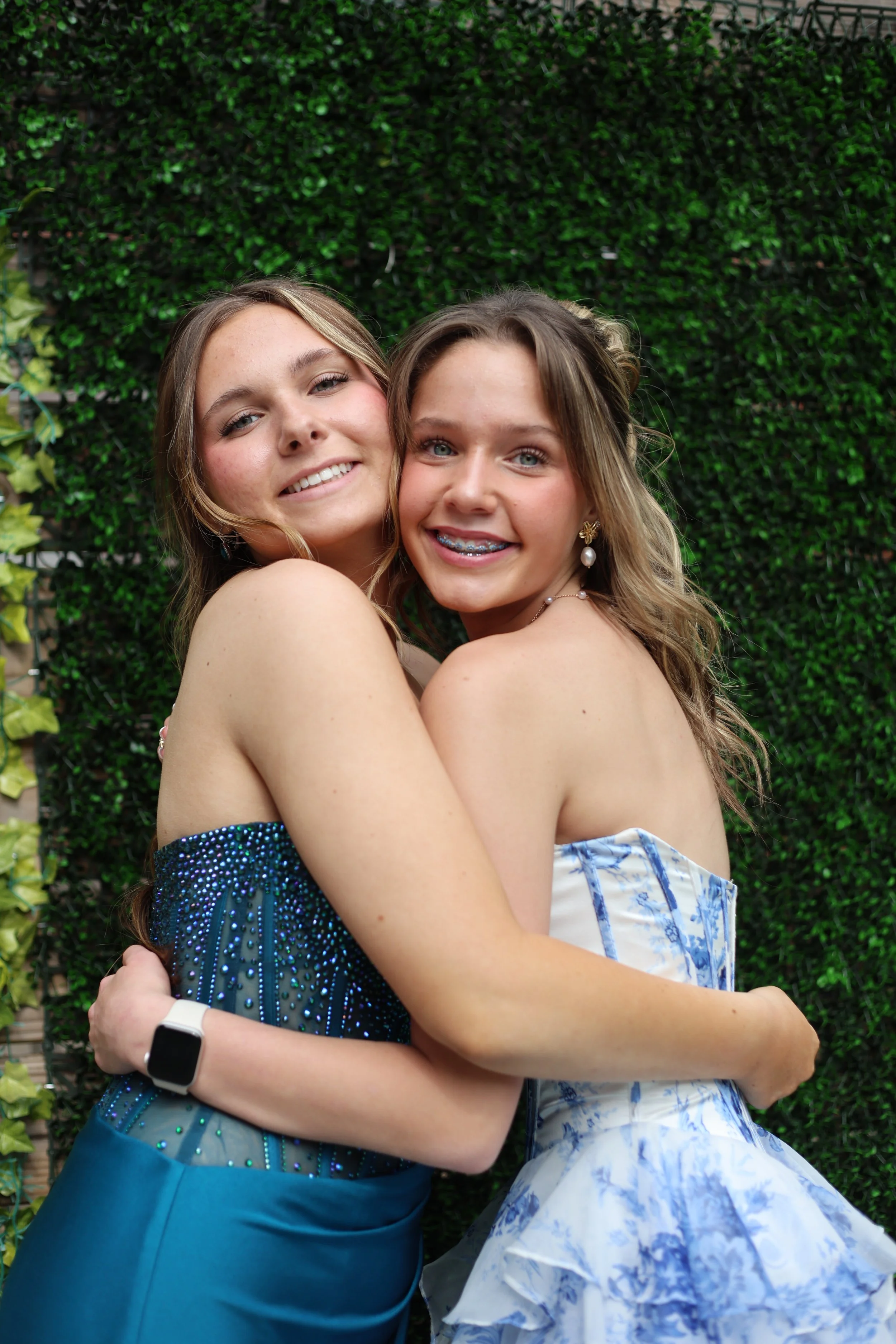 Two young women hugging each other and smiling in front of a green hedge backdrop.