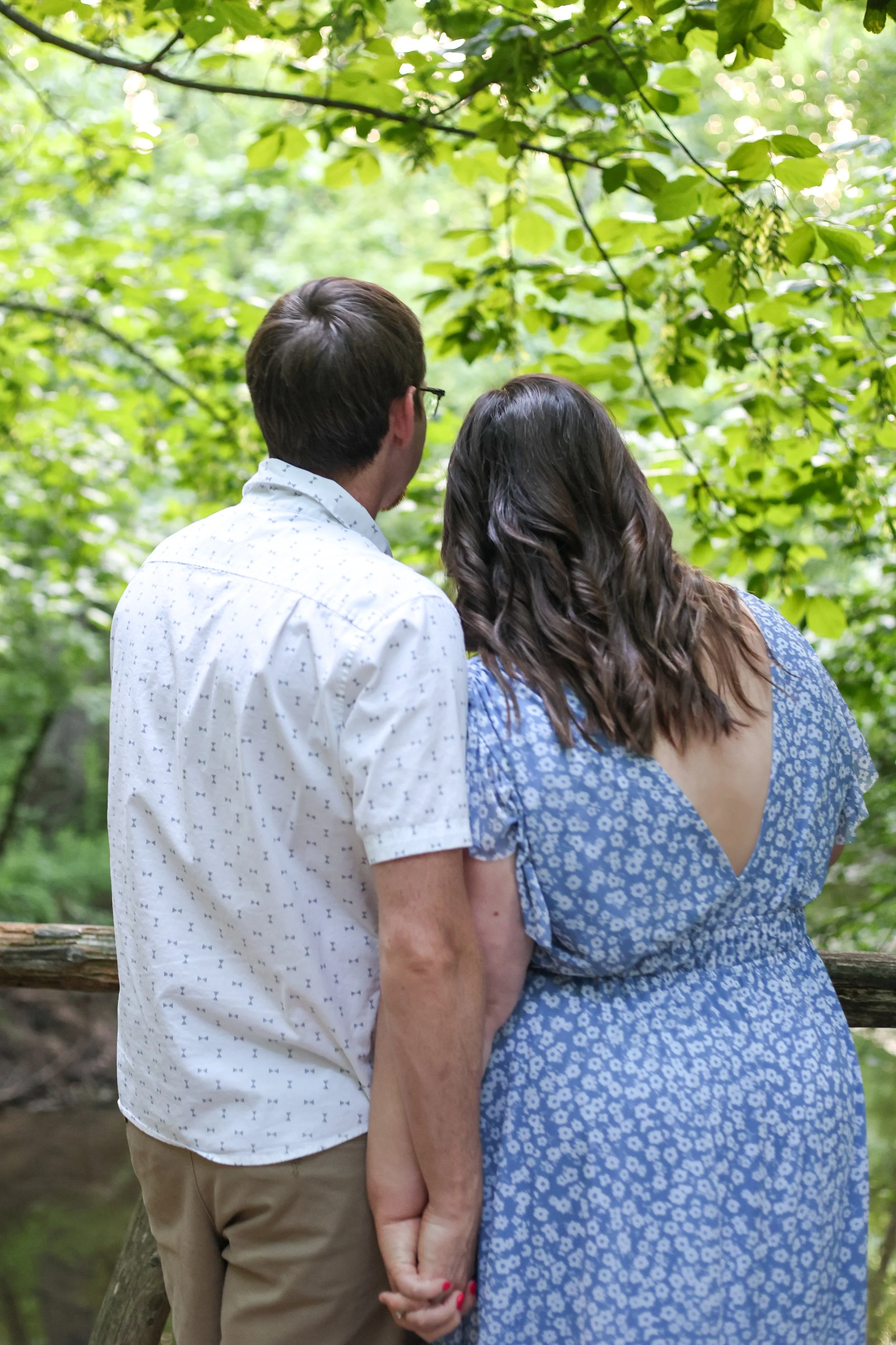 A couple holding hands stands together outdoors in a green forest, with their backs to the camera.
