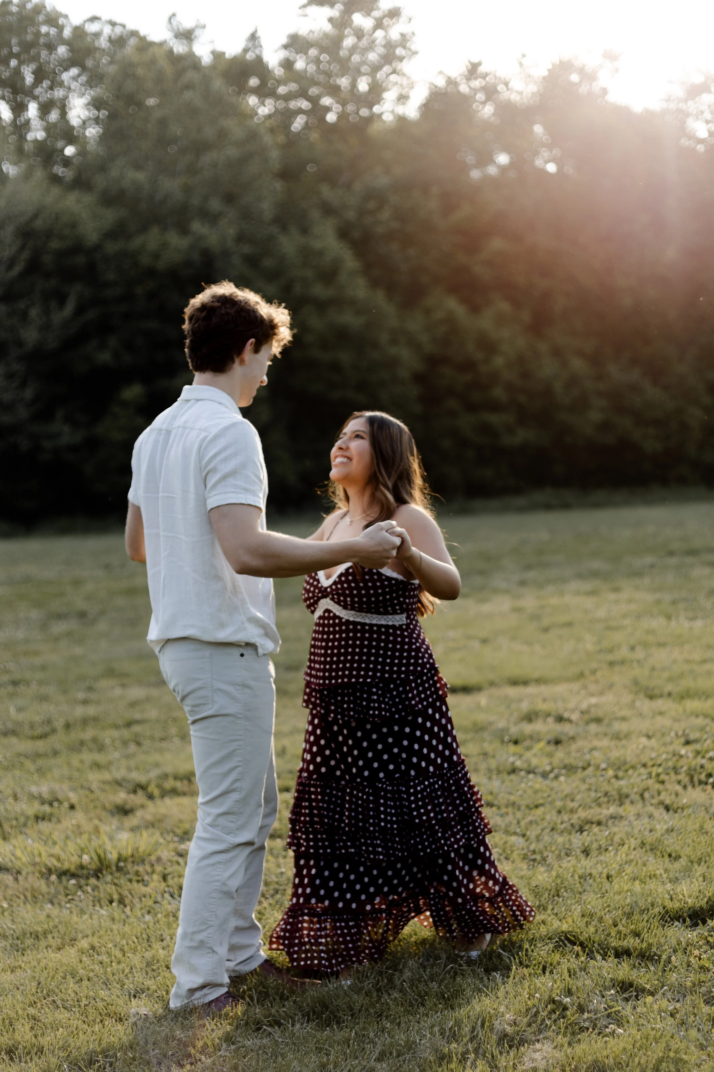 A young couple dancing outdoors on a grassy field during sunset with trees in the background.