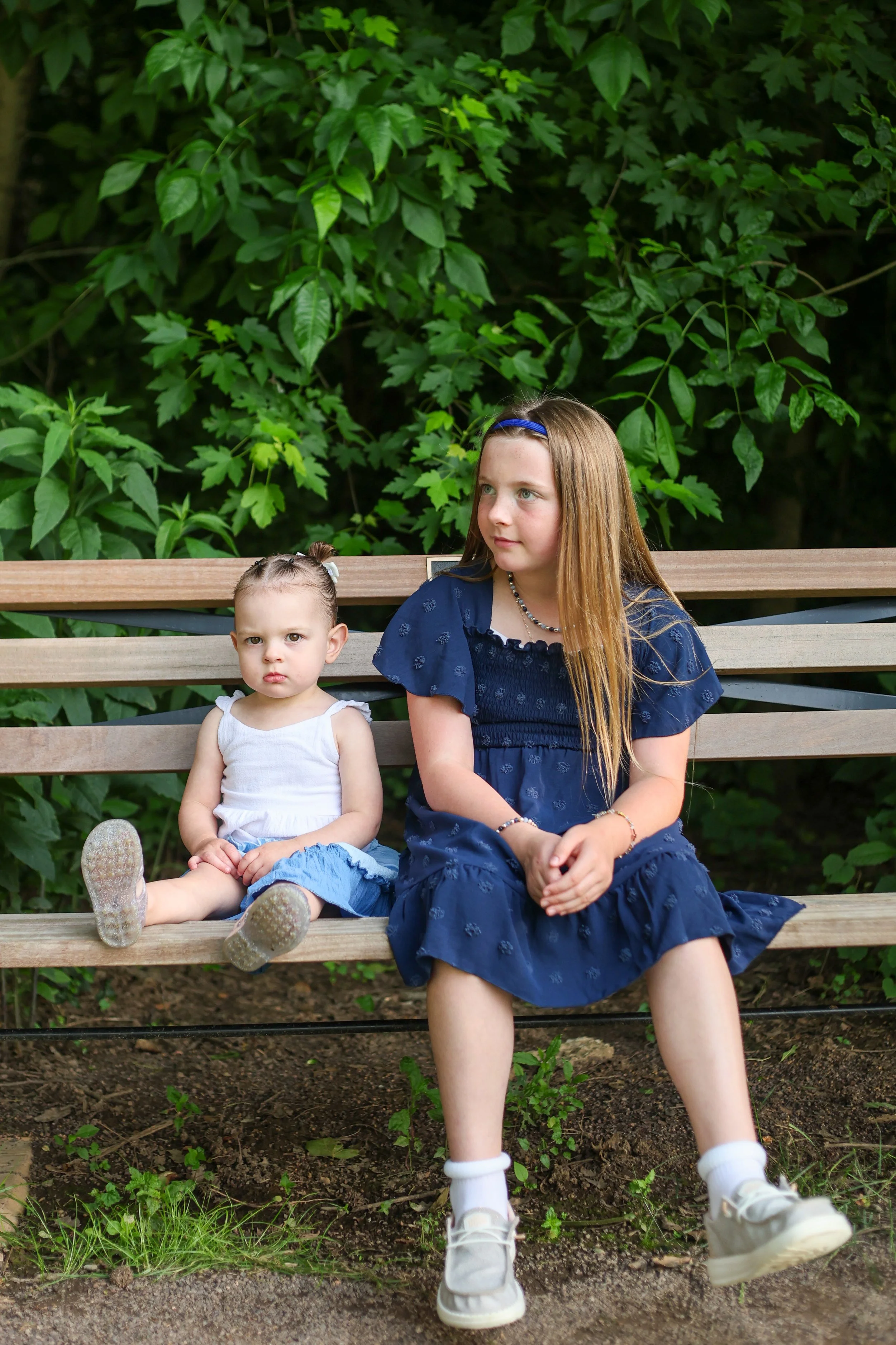 Two young girls sitting on a wooden park bench surrounded by green leafy trees. The younger girl, on the left, has light skin, brown hair in pigtails, and is wearing a white tank top and blue jeans. The older girl, on the right, has light skin, long 