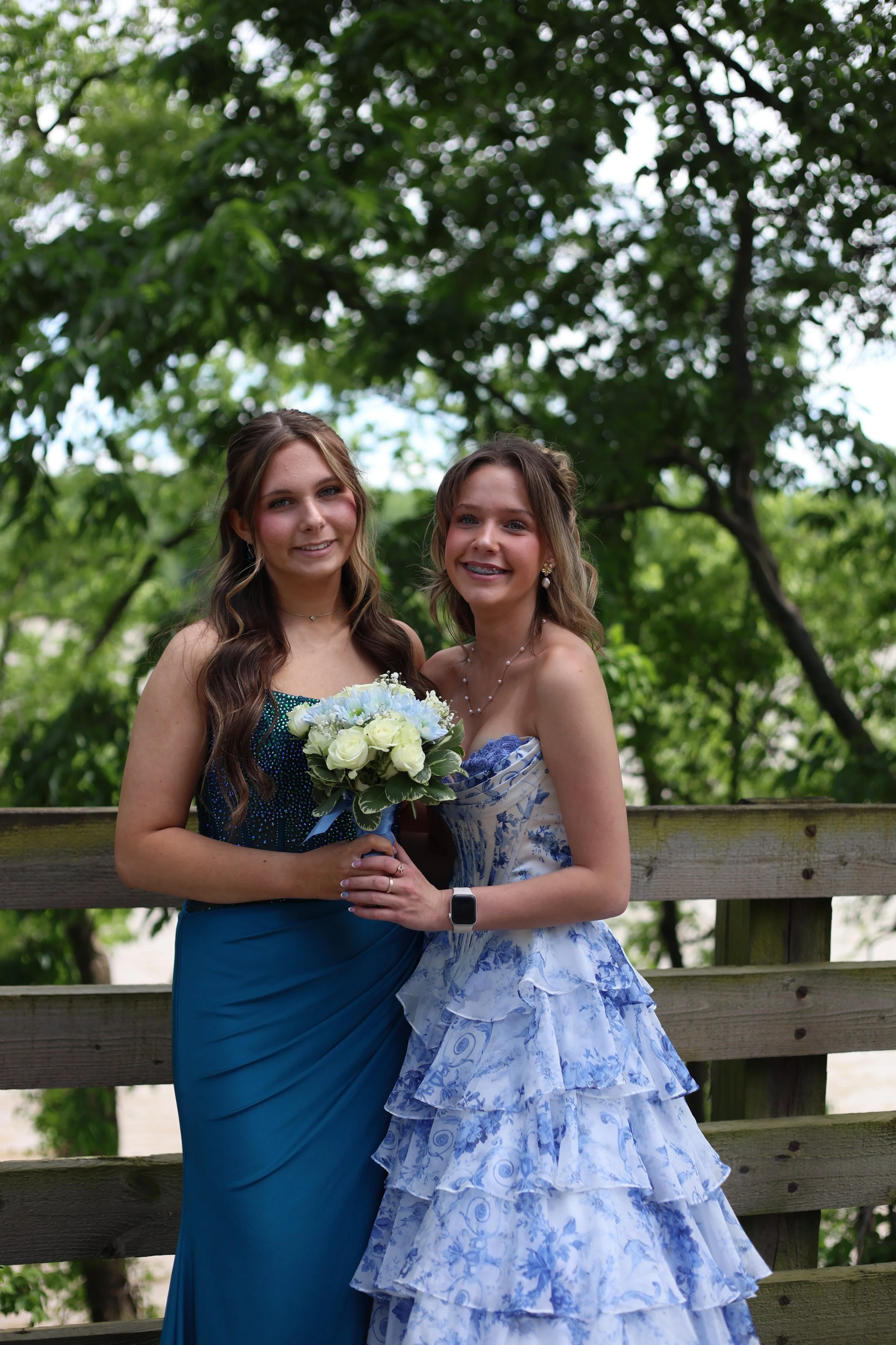 Two young women standing outdoors, dressed in formal attire, holding a bouquet of white roses. They are smiling and standing close together in front of a wooden fence with green trees in the background.