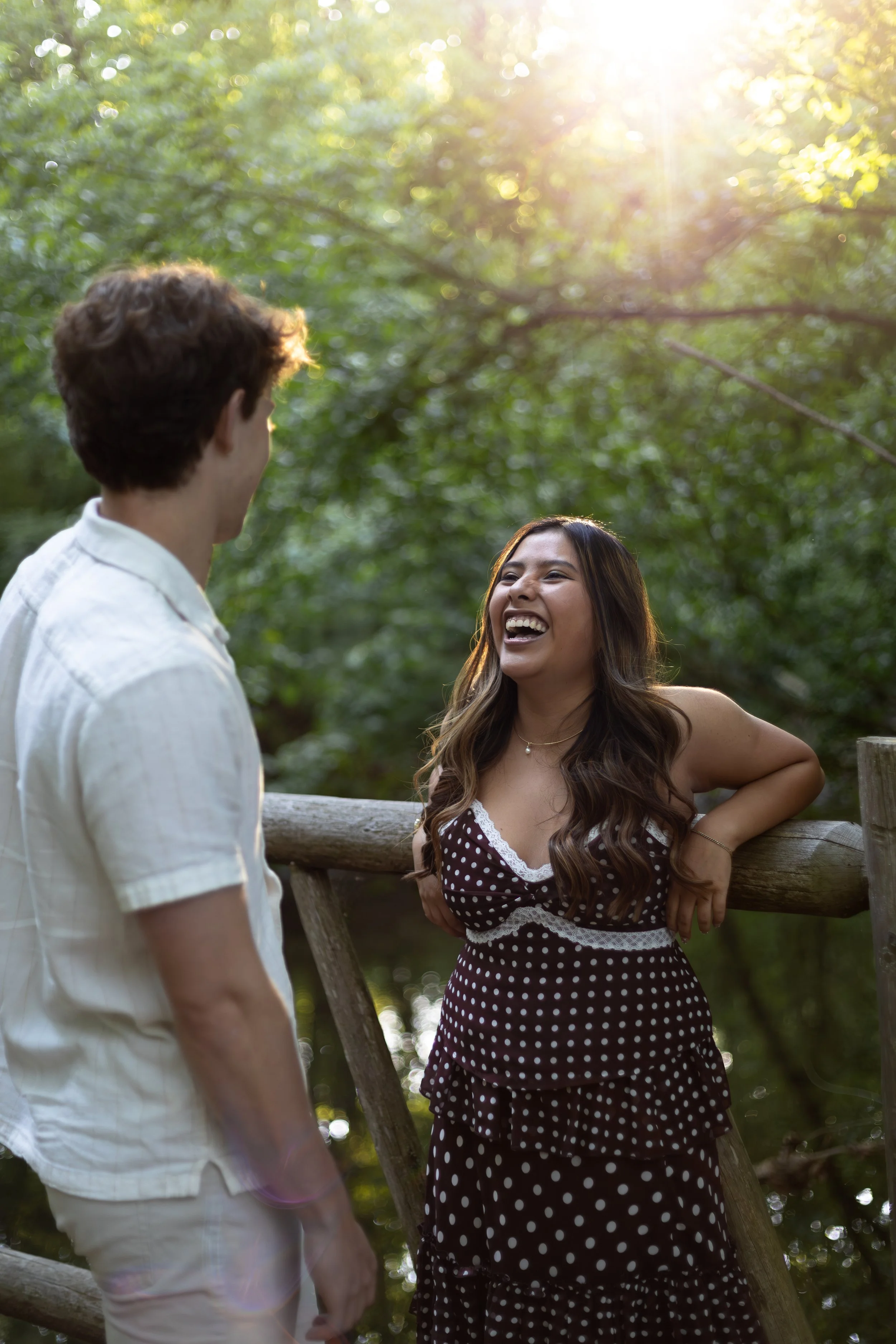 A man and a woman are standing outdoors on a wooden bridge, smiling and talking to each other. The woman has long, wavy hair and is wearing a polka dot dress, while the man has short, curly hair and is wearing a white shirt. Sunlight filters through 