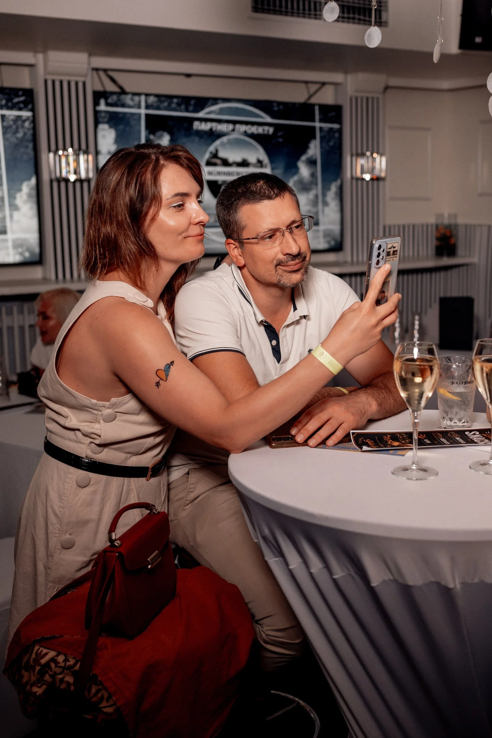 Ein Mann und eine Frau sitzen an einem runden Tisch, der Mann zeigt dem Bildschirm seines Handys, während die Frau zuschaut. Im Hintergrund eine Leinwand mit einem Wolkenbild und Text. Es ist eine Indoor-Event- oder Party-Atmosphäre.