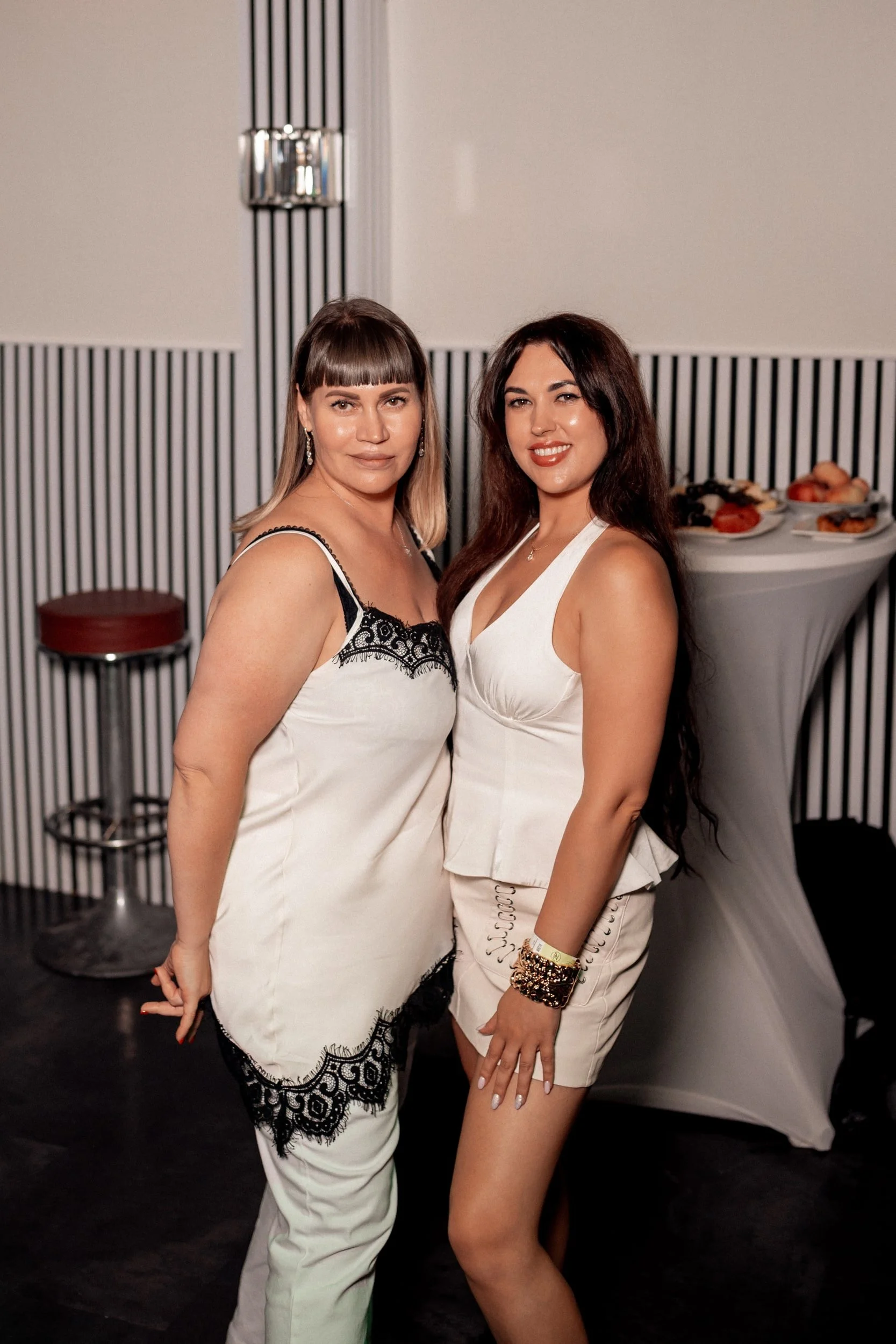 Zwei Frauen in Abendkleidern posieren bei einer Party, mit Finger-Pose, im Hintergrund ein Stehtisch mit Snacks.