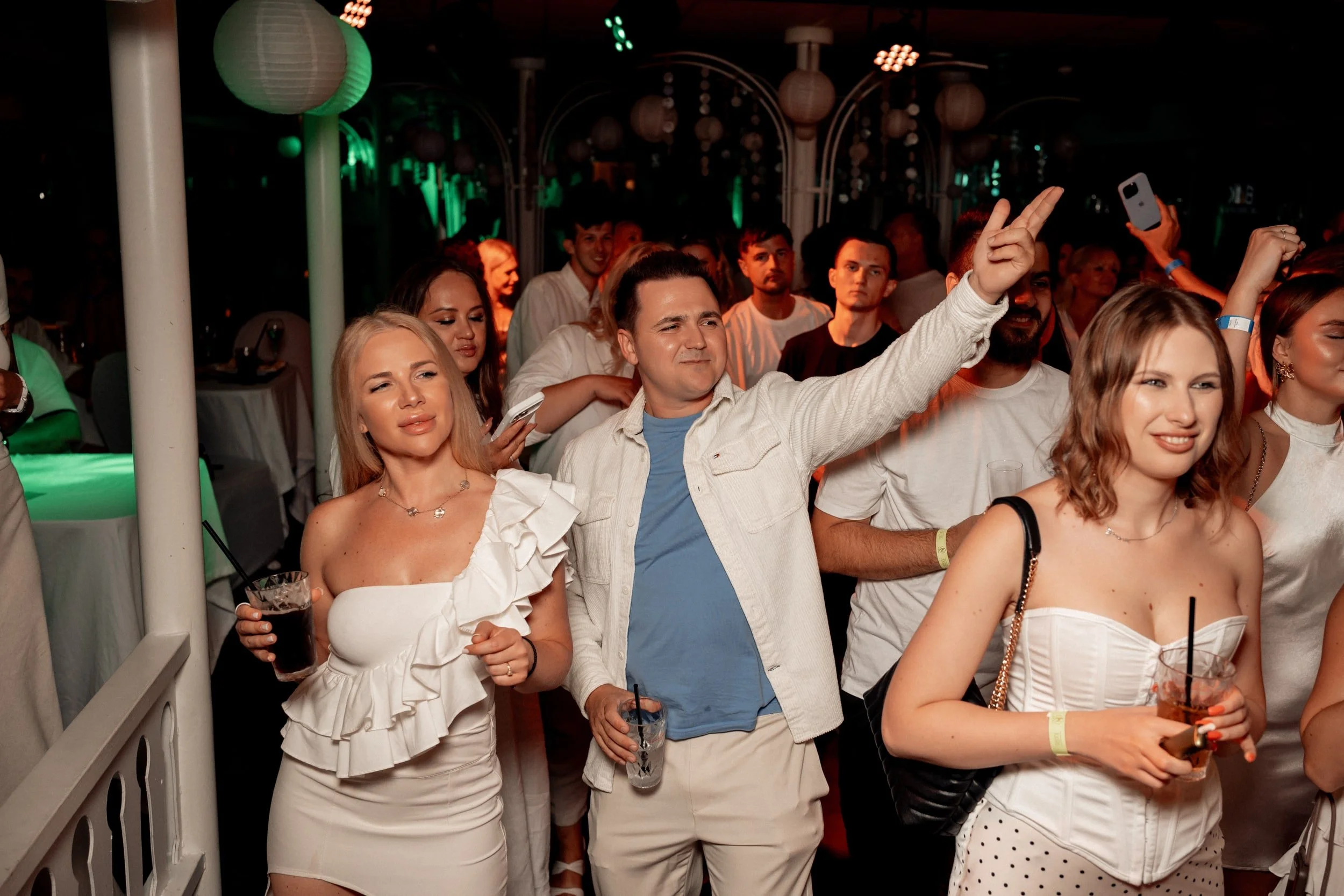 Menschen feiern auf einer Party im dunklen Raum mit bunten Lichteffekten, einige halten Getränke, eine Person macht ein Foto mit dem Handy.