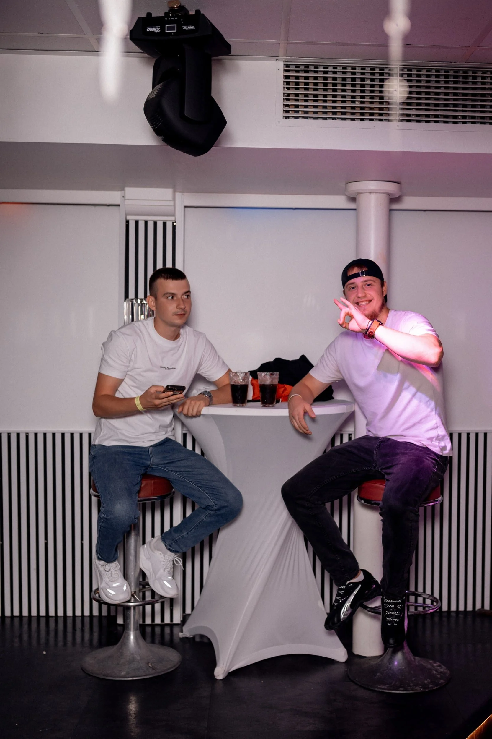 Zwei junge Männer sitzen an einem weißen Stehtisch in einer Bar oder einem Club. Der Mann auf der linken Seite trägt ein weißes T-Shirt und Jeans, während der Mann auf der rechten Seite ein weißes T-Shirt, schwarze Hose und einen schwarzen Baseballcap trägt. Beide haben Getränke vor sich, der Mann auf der linken Seite hält sein Smartphone. Der rechte Mann lächelt und macht eine Geste mit der Hand. Es gibt eine breite weiße Wand im Hintergrund, eine Säule und eine Deckenluftröhre. Eine große schwarze Lautsprecherbox hängt an der Decke.
