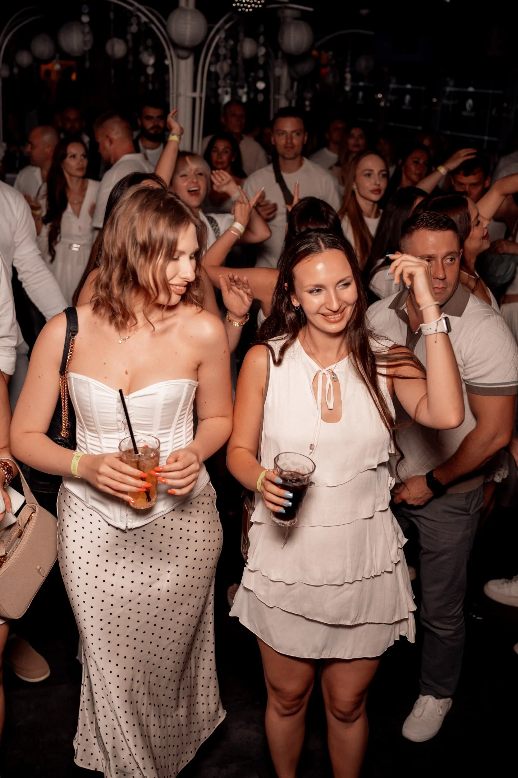 Menschen feiern auf einer Party, manche tanzen, einige halten Getränke, die Stimmung ist fröhlich, im Hintergrund sind laternenartige Dekorationen sichtbar.