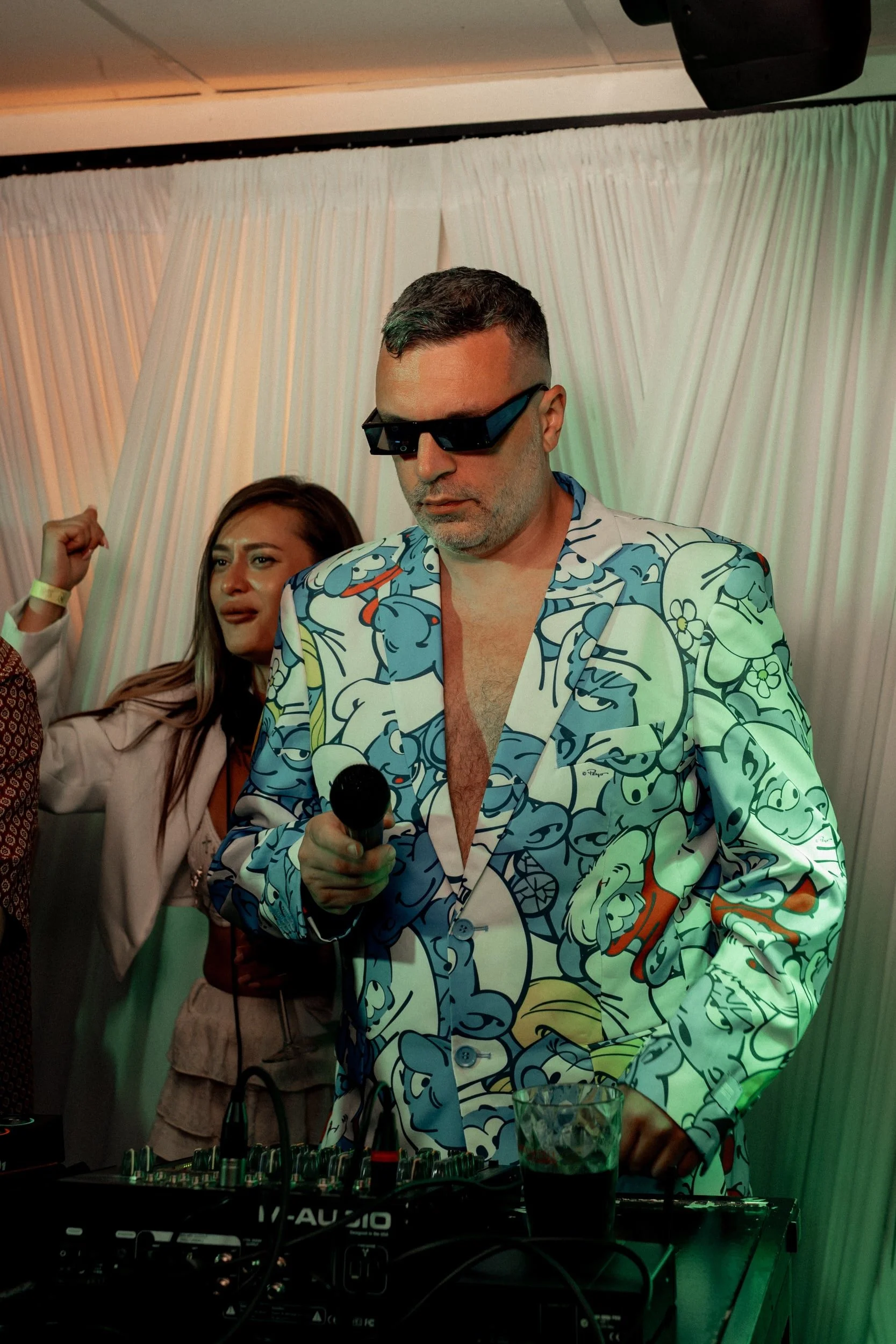 Ein Mann trägt ein bunt gemustertes Hemd mit Cartoon-Prints, schwarze Sonnenbrille, hält ein Mikrofon in der Hand und steht vor einem DJ-Pult. Eine Frau im Hintergrund, die ihre Faust in die Luft streckt, ist ebenfalls im Rahmen. Der Hintergrund hat weiße Vorhänge, die das Ambiente eines Events oder einer Party unterstreichen.