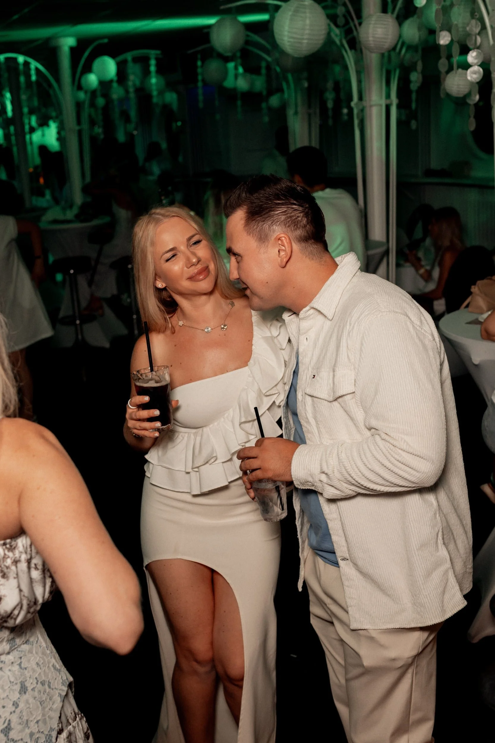 Ein Mann und eine Frau, die sich bei einer Feier oder Party nahekommen, mit dem Mann der die Frau küsst und diese lächelt. Die Frau trägt ein weißes Kleid mit offener Schulter und hält ein Glas mit Getränk. Das Bild ist in einem dunklen, festlichen Ambiente mit grüner Beleuchtung im Hintergrund.
