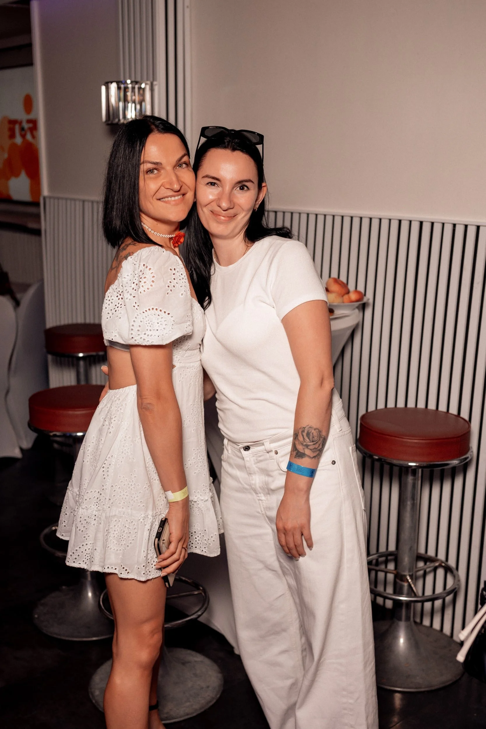 Zwei Frauen stehen lächelnd nebeneinander in einer Bar, eine trägt ein weißes Sommerkleid mit Löchern, die andere eine weiße Hose und T-Shirt. Im Hintergrund befinden sich Barhocker, Tische und eine Obstschale mit Äpfeln.