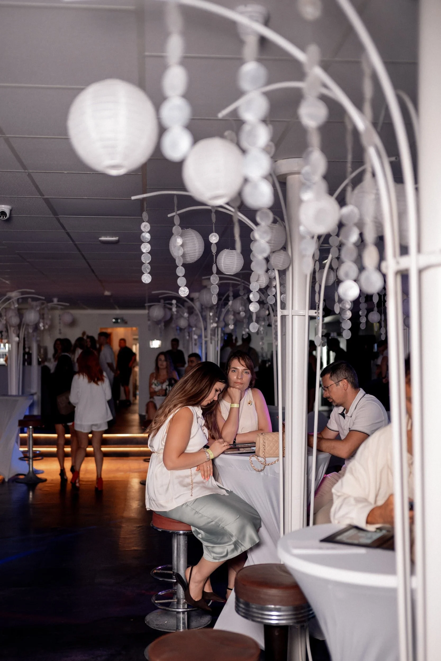 Menschen sitzen an einem Bar- oder Thekenbereich, während im Hintergrund Gäste im Aufnahmeraum stehen. Die Decke ist verziert mit weißen, leuchtenden Lampions, die wie Papierlaternen aussehen.
