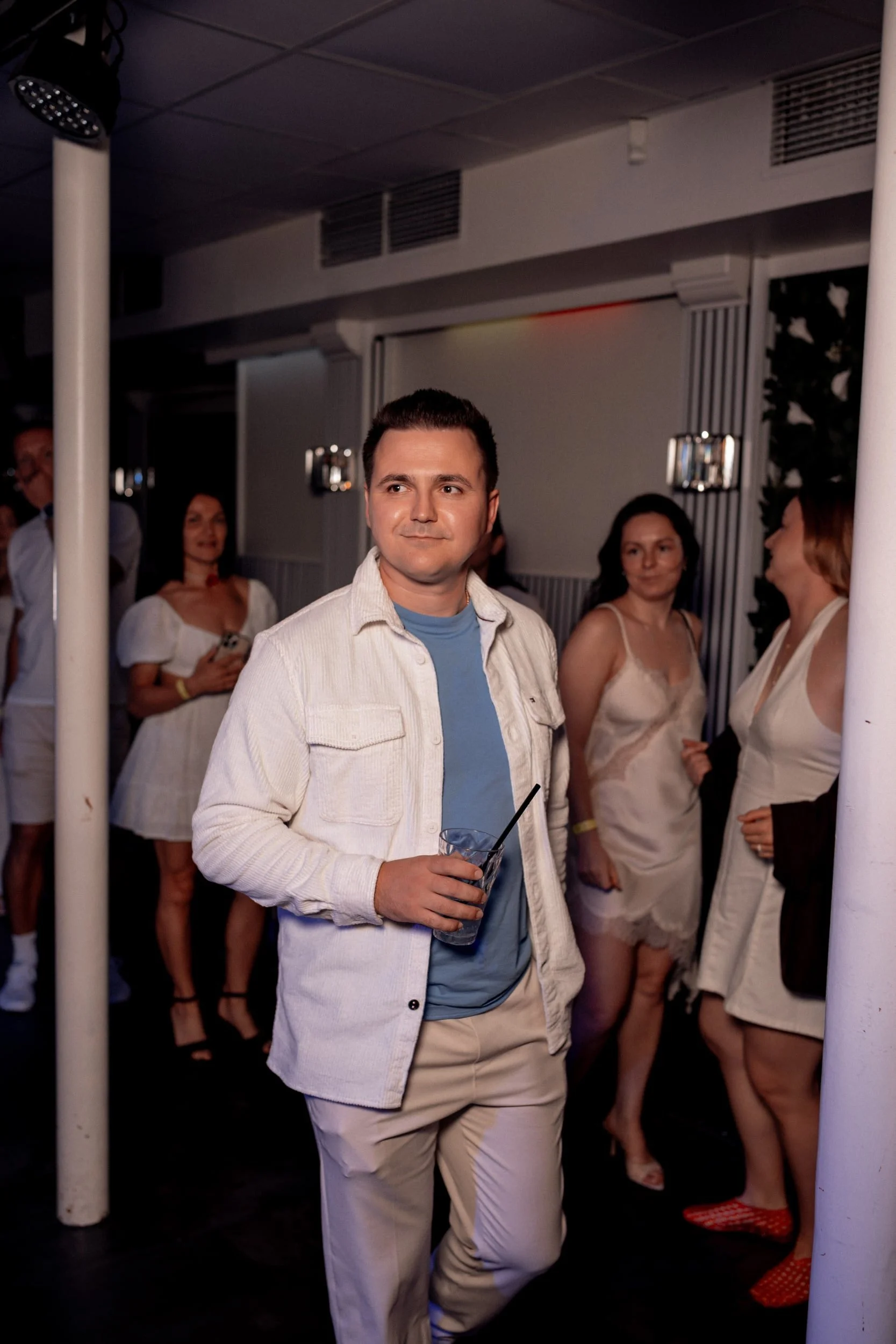 Ein junger Mann mit dunklem Haar trägt ein helles Jackett und hält ein Getränk in der Hand, während er in einer Gruppe von Frauen auf einer Party steht.