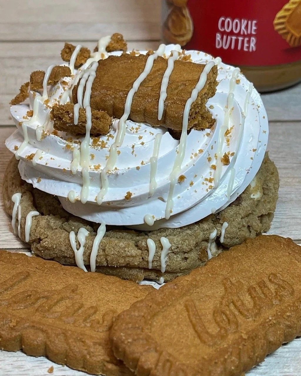A cookie topped with marshmallow fluff, cookie chunks, and white chocolate drizzle, surrounded by more cookies and a jar of cookie butter in the background.