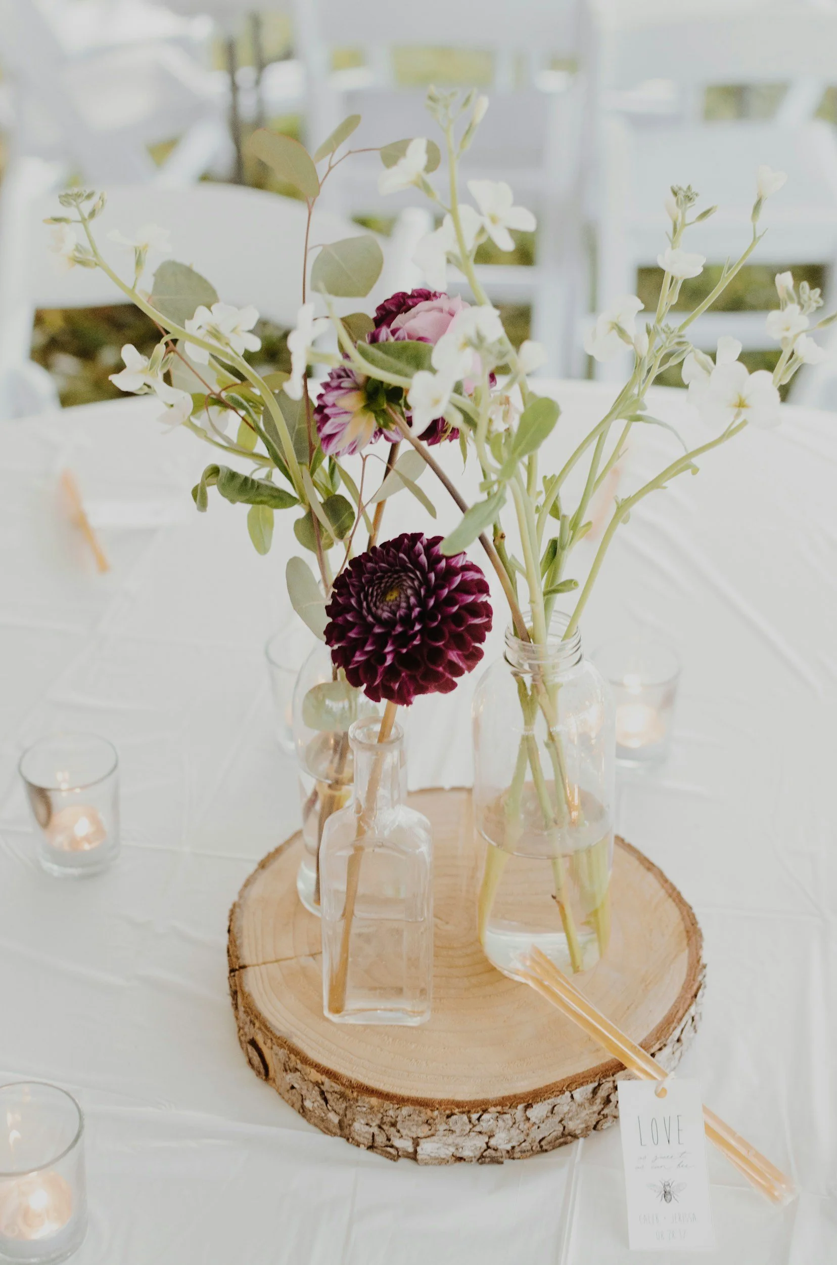 A floral centerpiece with pink and dark purple dahlias, white flowers, and green leaves in small glass bottles on a wooden slab, surrounded by tea light candles on a white tablecloth at an event.