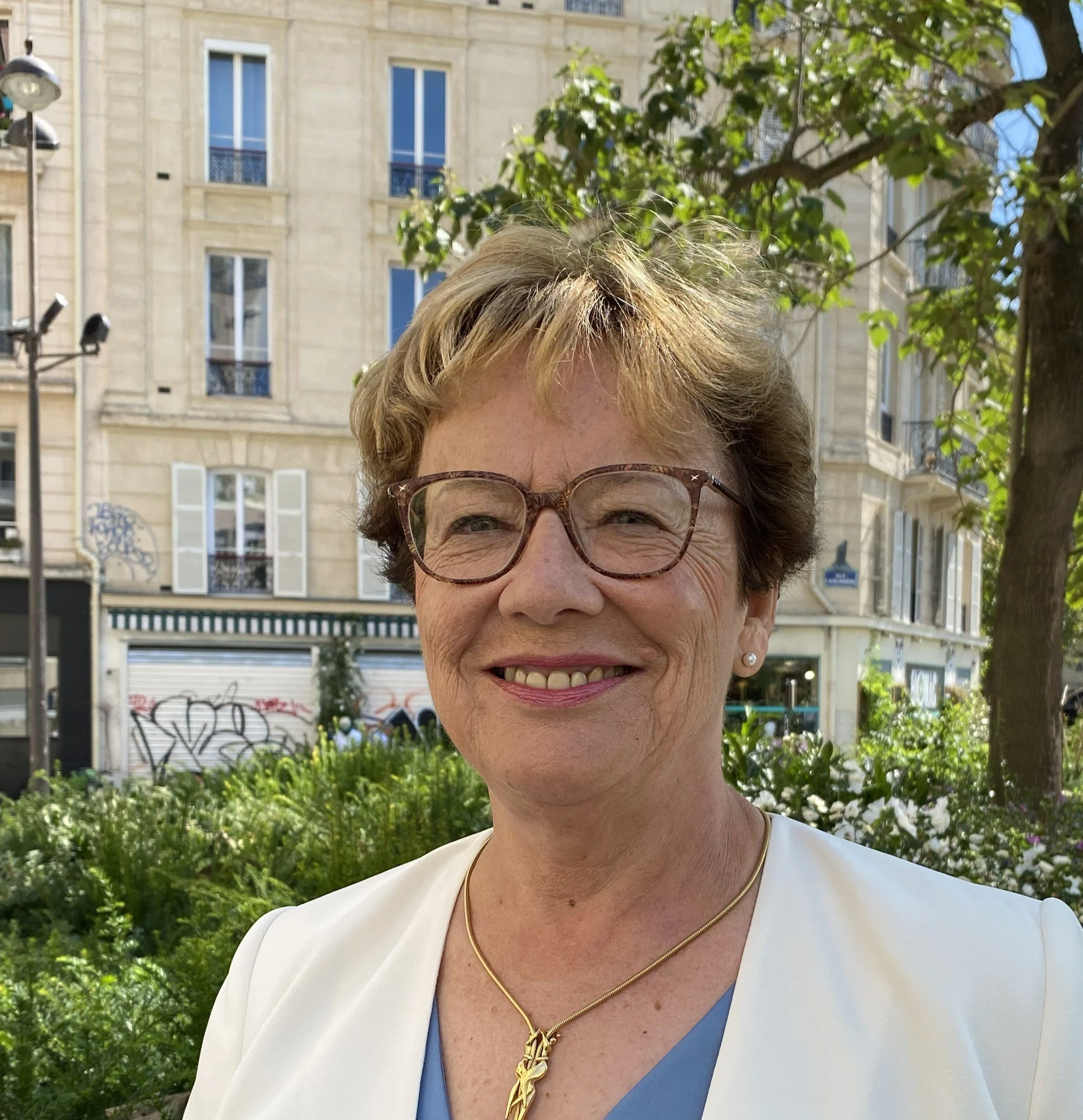 Une femme âgée souriante portant des lunettes, un collier en or et une veste blanche, à l'extérieur dans un quartier résidentiel avec des bâtiments en pierre et des arbres.