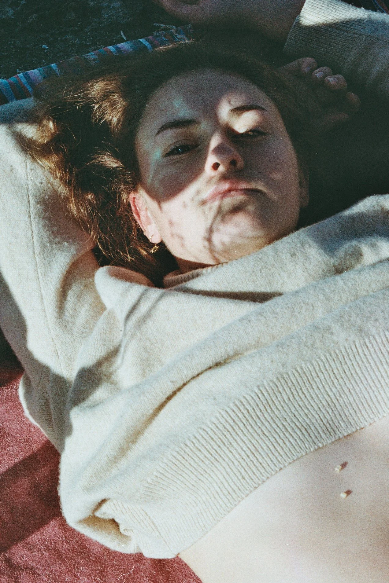 Une jeune femme à la peau claire, avec des cheveux courts bruns bouclés, porte un pull beige et se trouve sur un tapis rose. Elle regarde l'appareil photo avec une expression neutre.