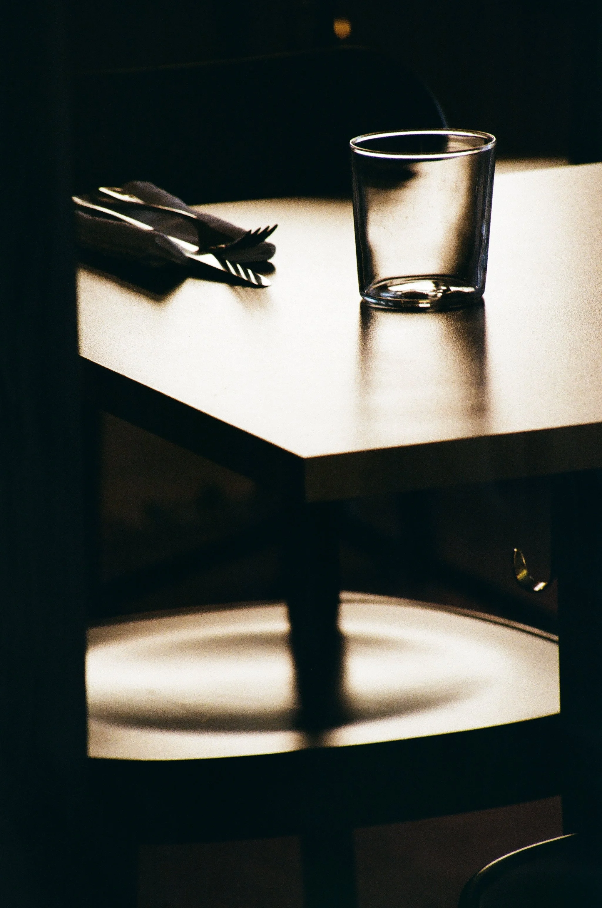 Photographie très contrastée ou la lumière en contre jour joue un rôle essentiel. Un gros plan d'un verre vide posé sur une table dans un restaurant, avec une cuillère et une fourchette posées à côté.
