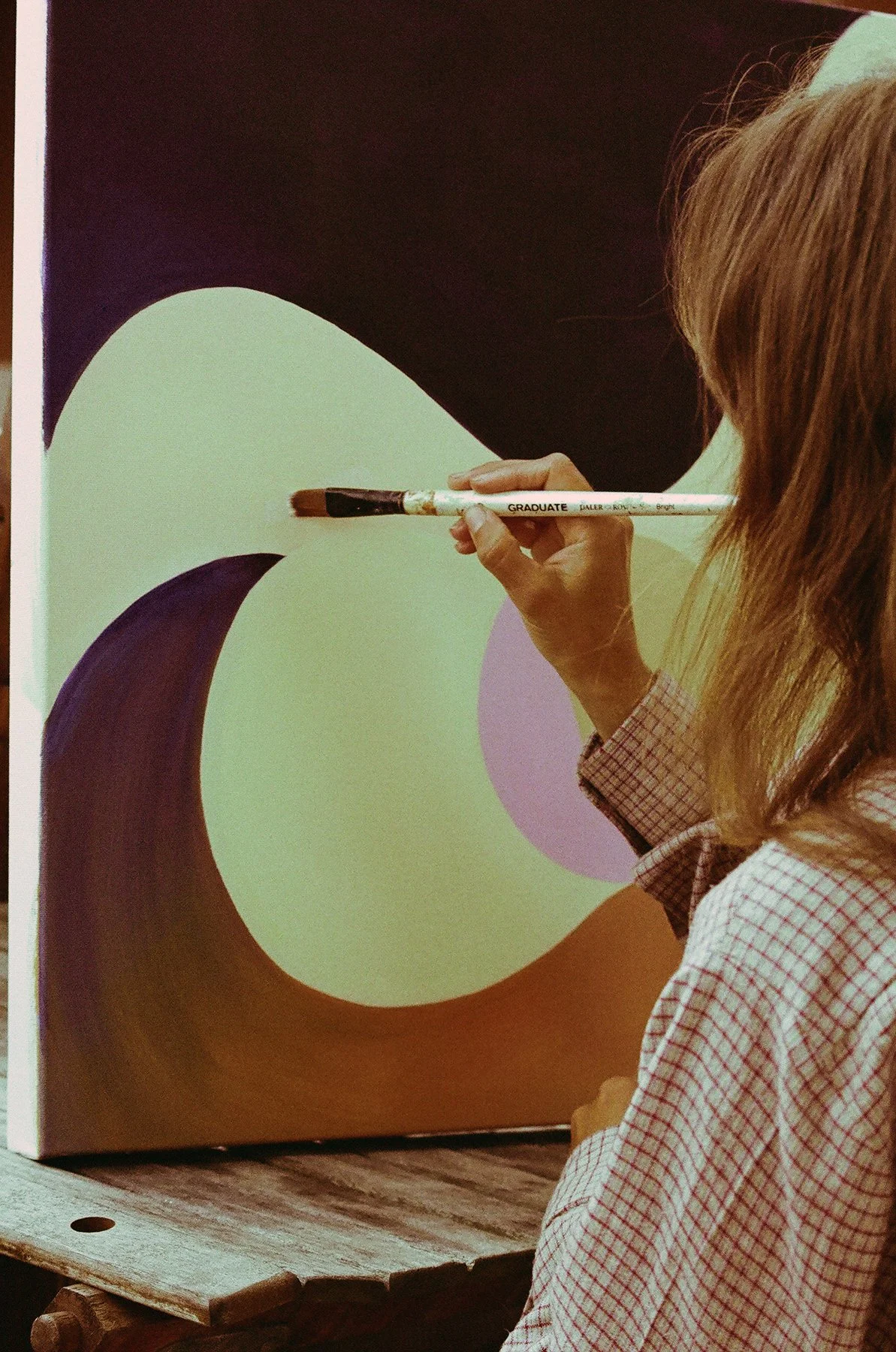 Une personne en train de peindre un tableau abstrait avec des formes courbes et des couleurs pastel, utilisant un pinceau large.