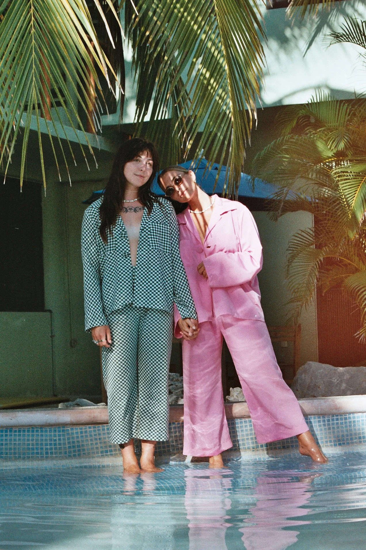 Deux femmes debout dans une piscine avec des palmiers en arrière-plan, tenant la main, portant des vêtements colorés et des accessoires de mode.
