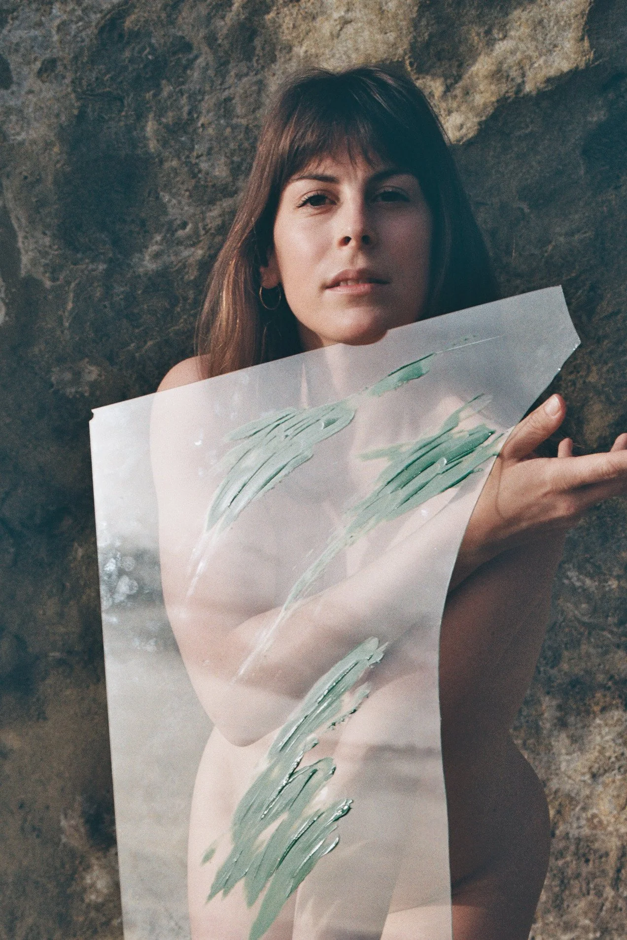 Une femme avec des cheveux bruns et un regard fixe pose contre un mur rocheux, tenant une plaque en verre cassé avec des traits verts peints dessus.