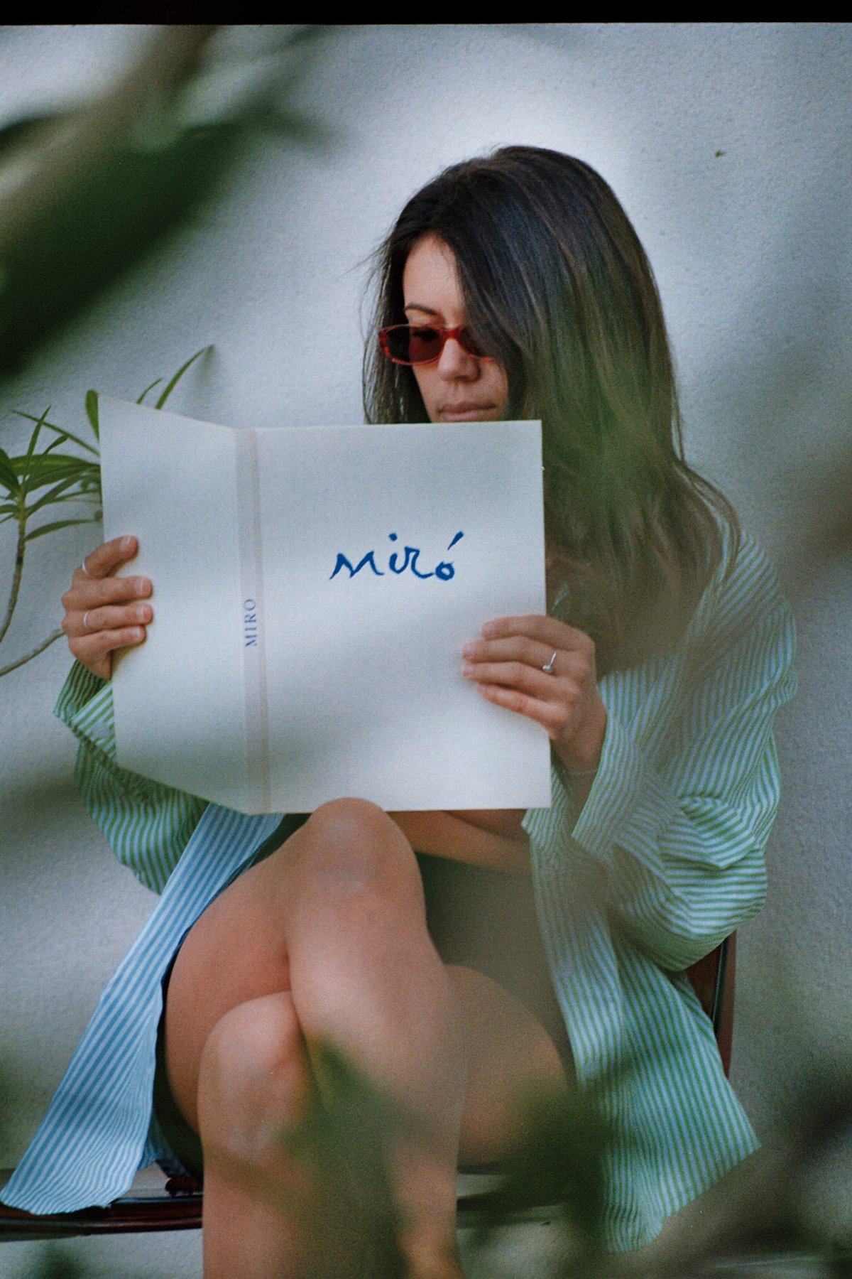 Une femme portant des lunettes de soleil rouges lit un livre blanc avec le mot "míró" écrit dessus. Elle est assise et porté une chemise à rayures. La photo est prise de près et inclut une partie de sa jambe au premier plan.
