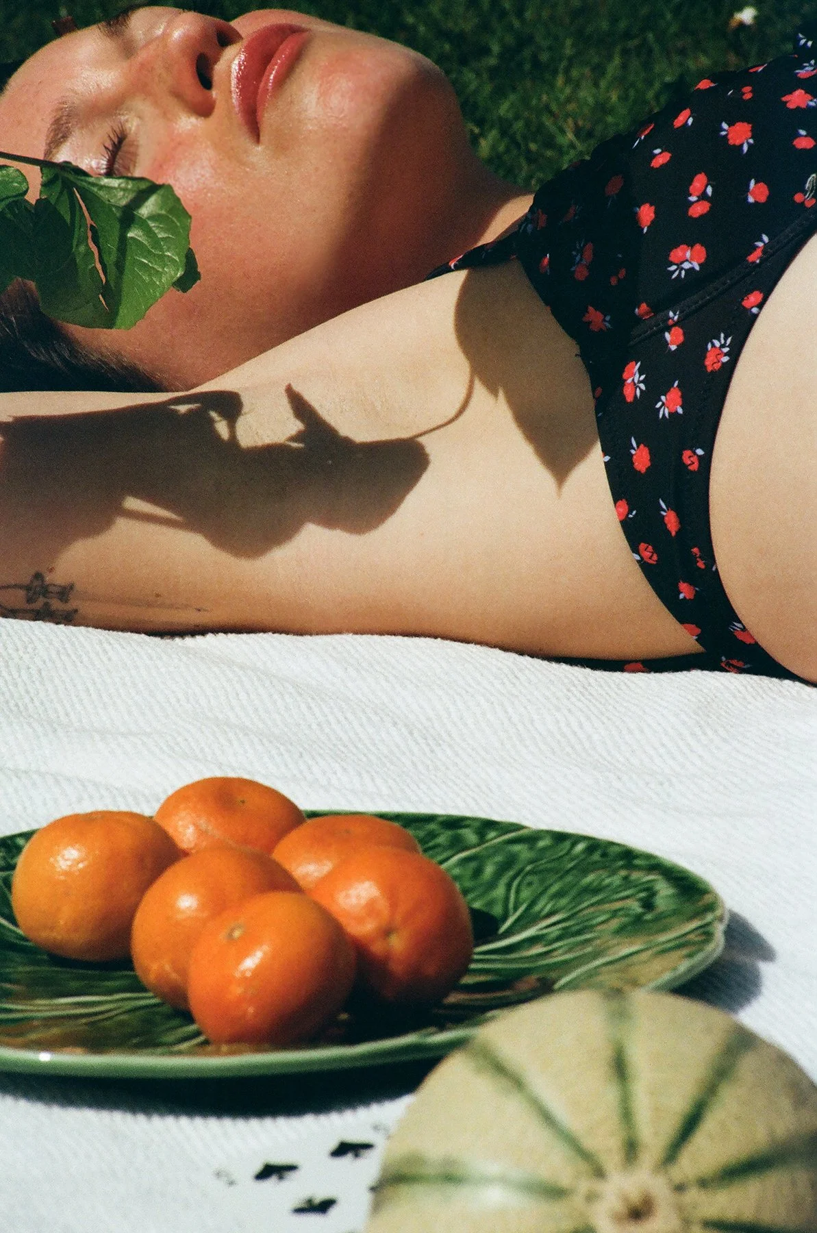 Femme allongée sur l'herbe portant une robe noire à petites fleurs rouges, ombragée partiellement par une feuille. Sur la table blanche devant elle, un plat vert avec des mandarines. En bas, partiellement visible, une feuille d'une plante
