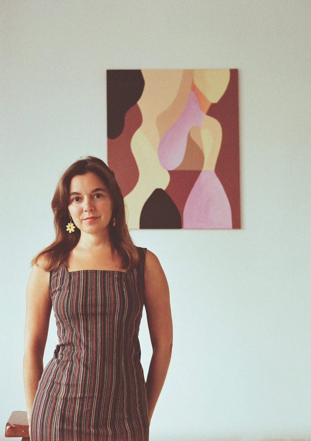Une femme portant une robe rayée se tient devant un tableau abstrait aux couleurs chaudes, dans une pièce avec murs blanc.