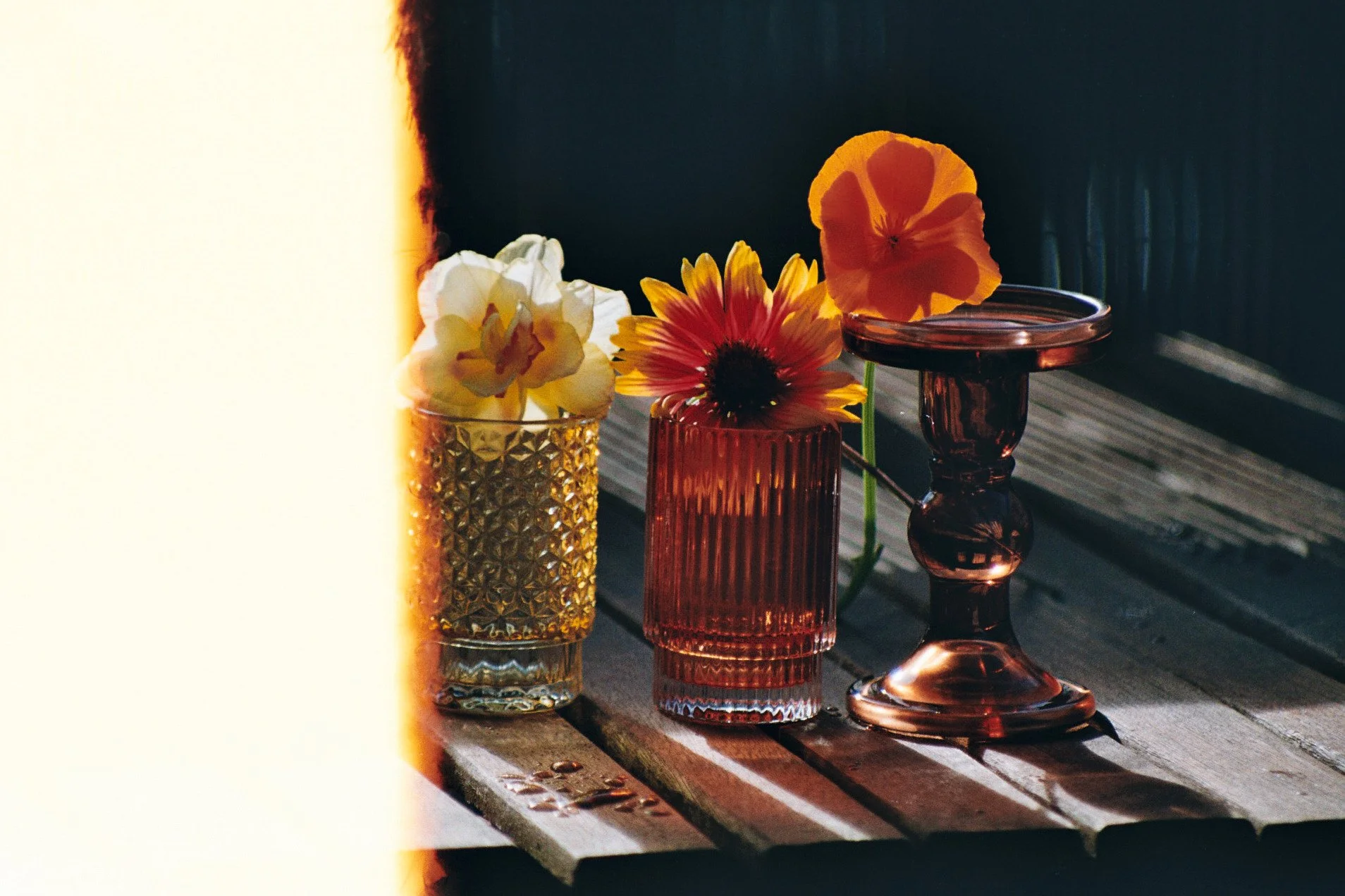 Trois vases de différentes couleurs contenant des fleurs colorées, posés sur une table d'exterieure en bois avec un fond sombre. La lumière naturelle éclaire partiellement la scène.