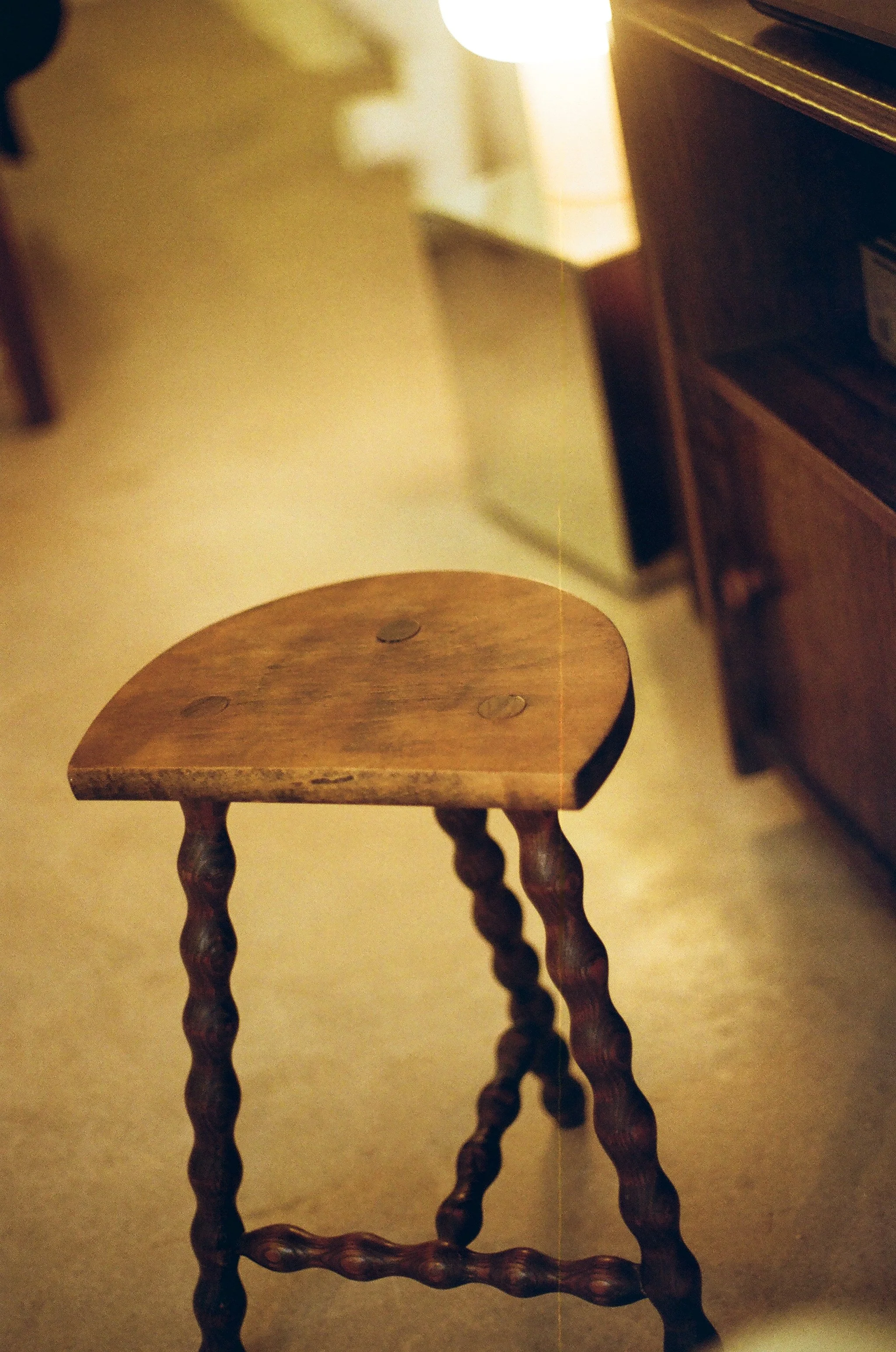 Une petite chaise en bois avec des pieds torsadés est au centre de l'image. En arrière-plan, un meuble en bois et un objet lumineux sont visibles.