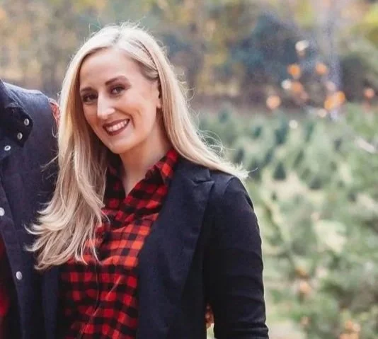 A smiling woman with long blonde hair, wearing a black jacket over a red and black plaid shirt, outdoors in a natural setting with trees and greenery in the background.