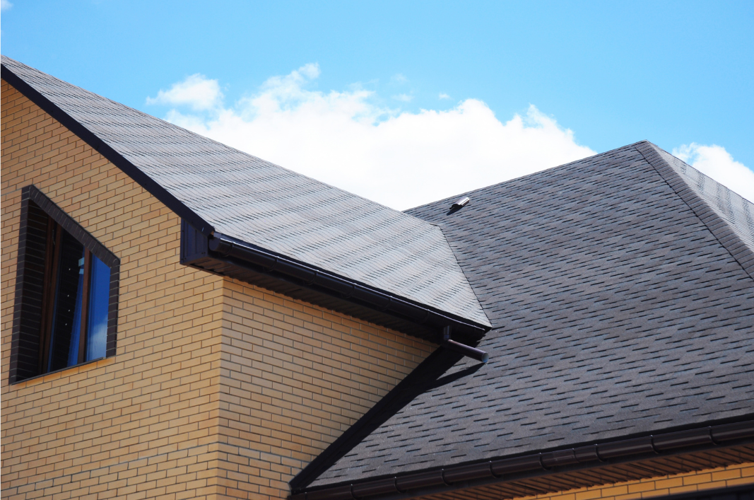 Close-up of a house's roof with dark shingles and a brick exterior wall, under a blue sky with few clouds.