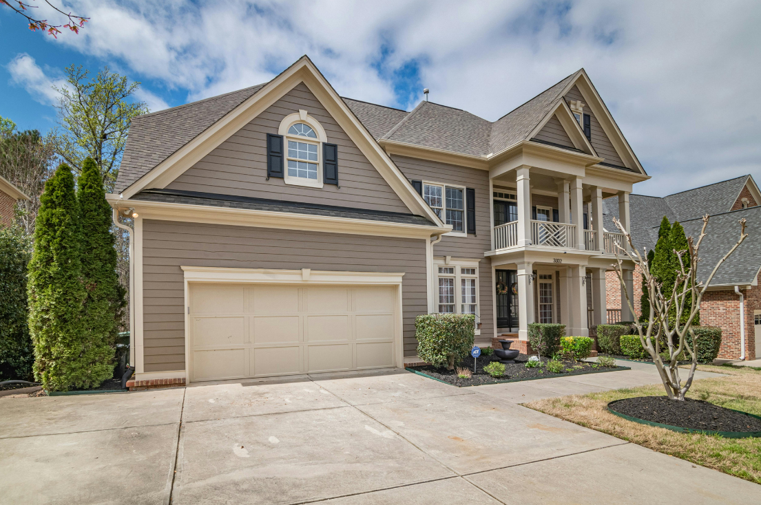 Front view of a two-story beige house with black shutters, a garage, and a small balcony, surrounded by a landscaped yard with bushes and a leafless tree.
