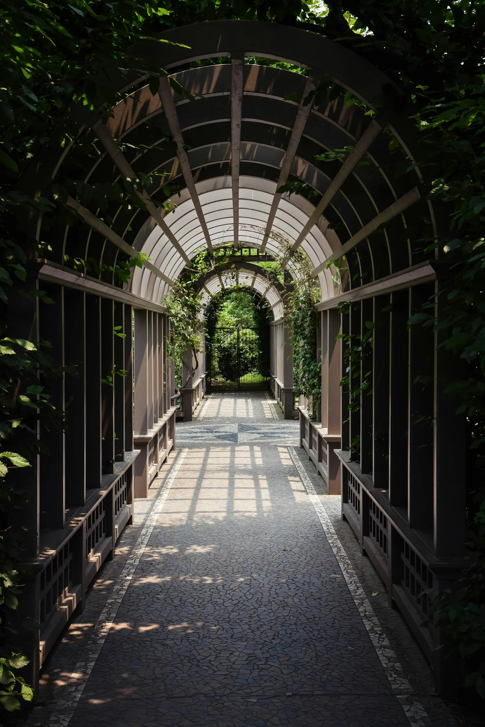 A covered pathway with an arched, metal and glass roof. Surrounding the pathway are green plants and trellises, with a tiled floor leading to a garden gate in the distance. Shades of sunlight create shadows on the ground.