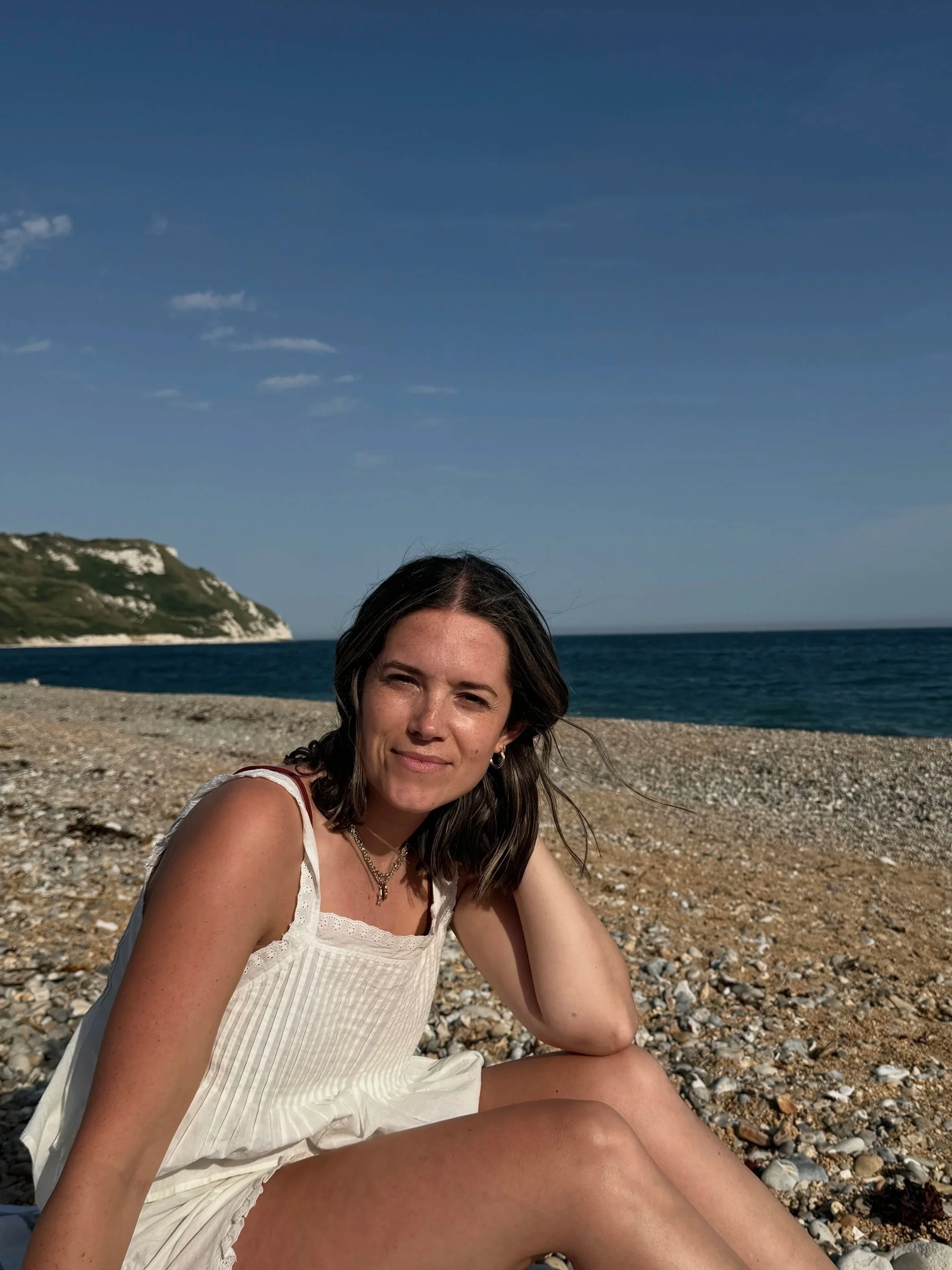 A woman with dark brown hair and earrings sitting on a rocky beach with the ocean and cliffs in the background, smiling slightly under a clear blue sky.