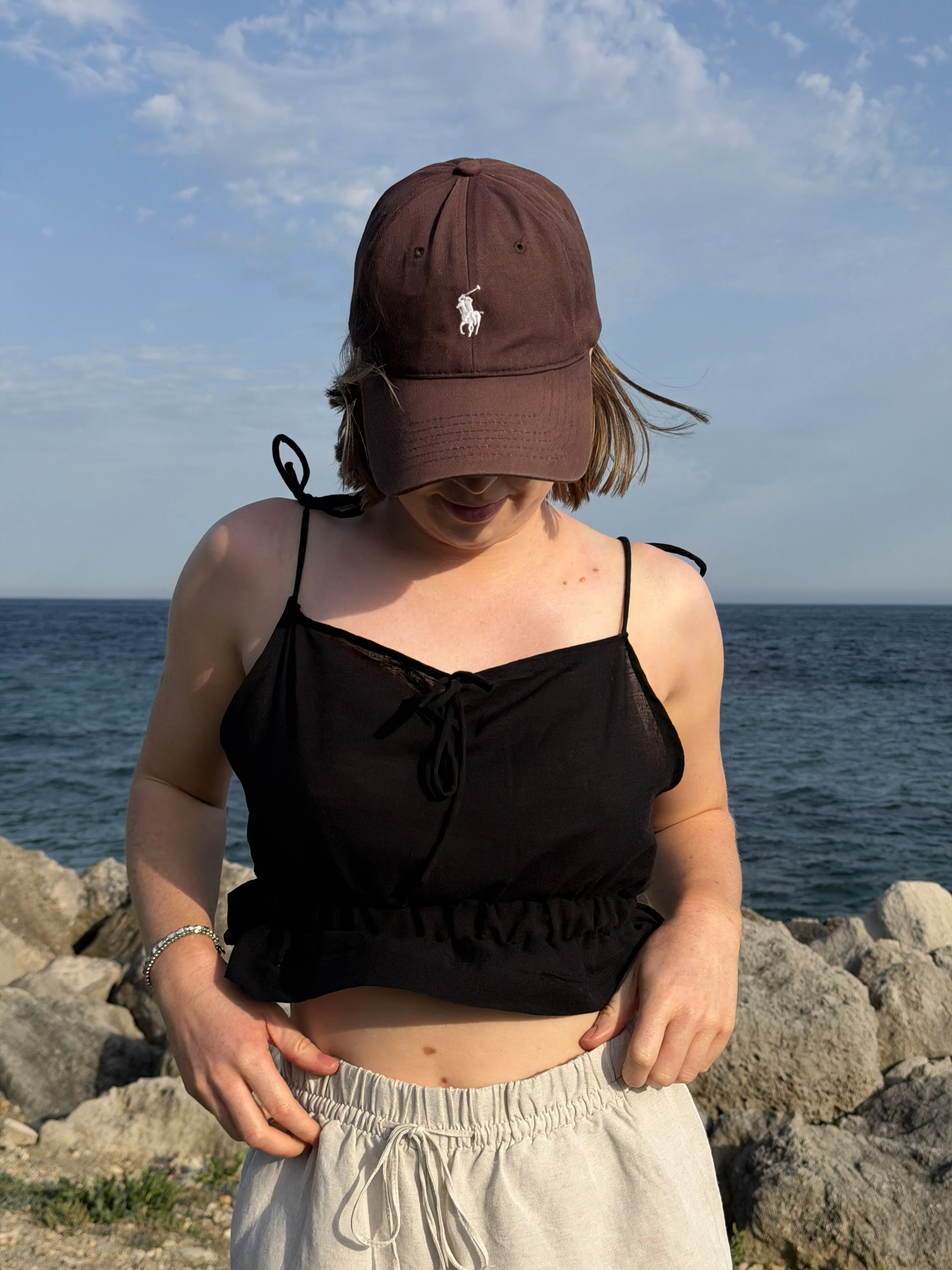 Content Capture for Herbs & Olive with a woman standing outdoors near the ocean, wearing a brown cap with a white logo and a black top, with her head tilted downward, and her hands resting on her waist, with rocks and the sea in the background.