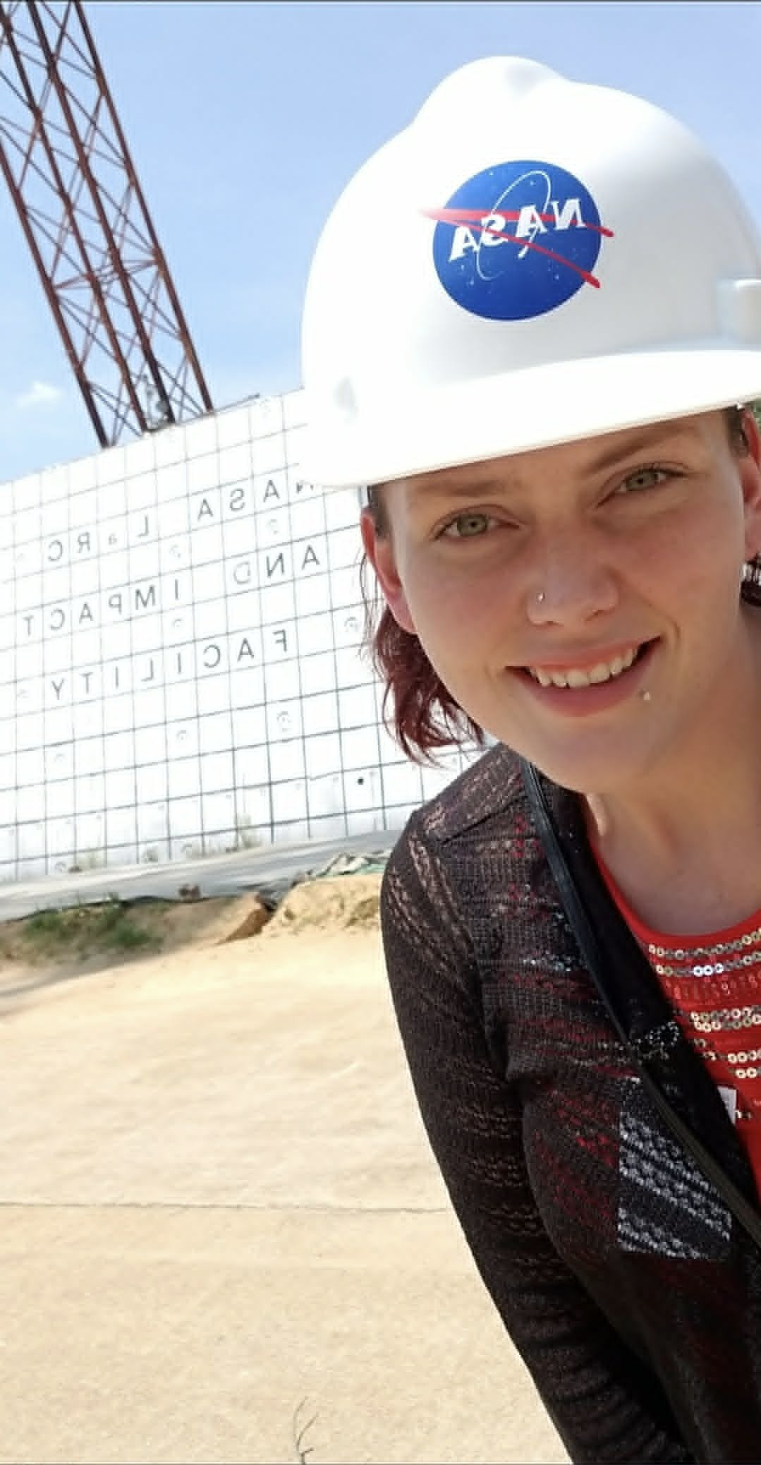 A woman with a nose piercing smiling at the camera, wearing a white hard hat with NASA logo, standing in front of a large white structure at a space launch site with a red and white tower in the background.