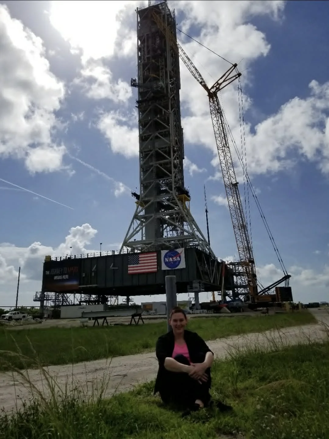 A woman sitting on grass in front of a large NASA rocket launch pad with a tall tower and cranes, and an American flag.