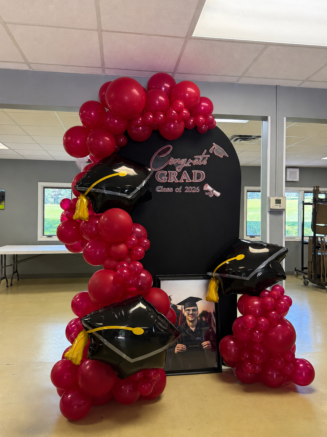 Graduation photo display with balloons, including black mortarboards and pink balloons, a framed photo of a graduate, and a sign reading 'Congrats Grad Class of 2026' with pink and black accents.