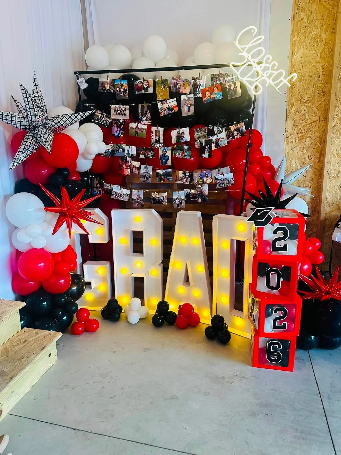 Decorative setup featuring a balloon arch with red, black, and white balloons, string lights, a large illuminated 'Grad' sign, photo display clips hanging on strings, star and graduation cap shapes, and a vertical stacking game with numbers from 0 to