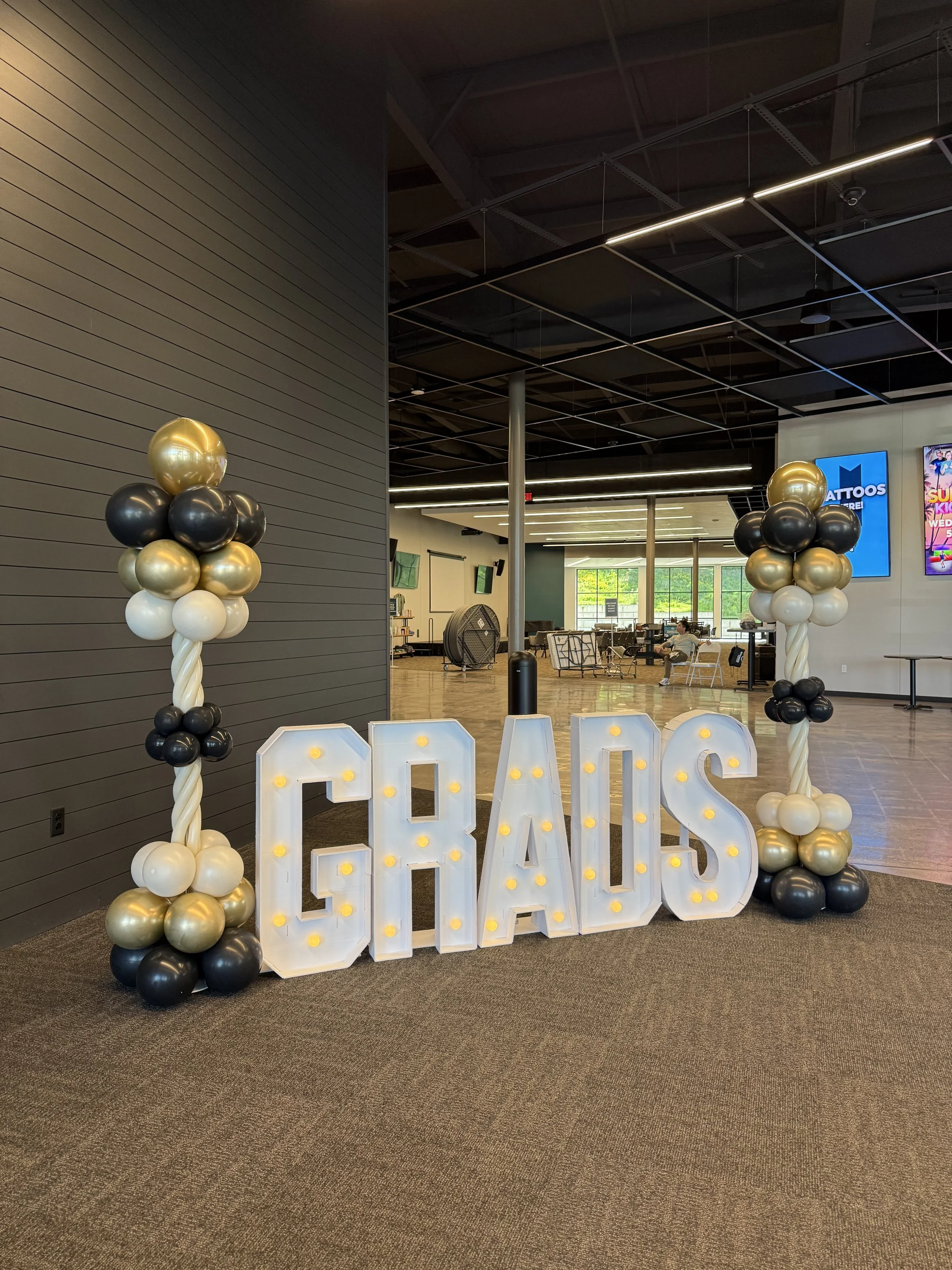 Decorative balloon display with gold, white, and black balloons, and a large illuminated sign spelling "GRADS" for graduation celebration in a modern indoor space.