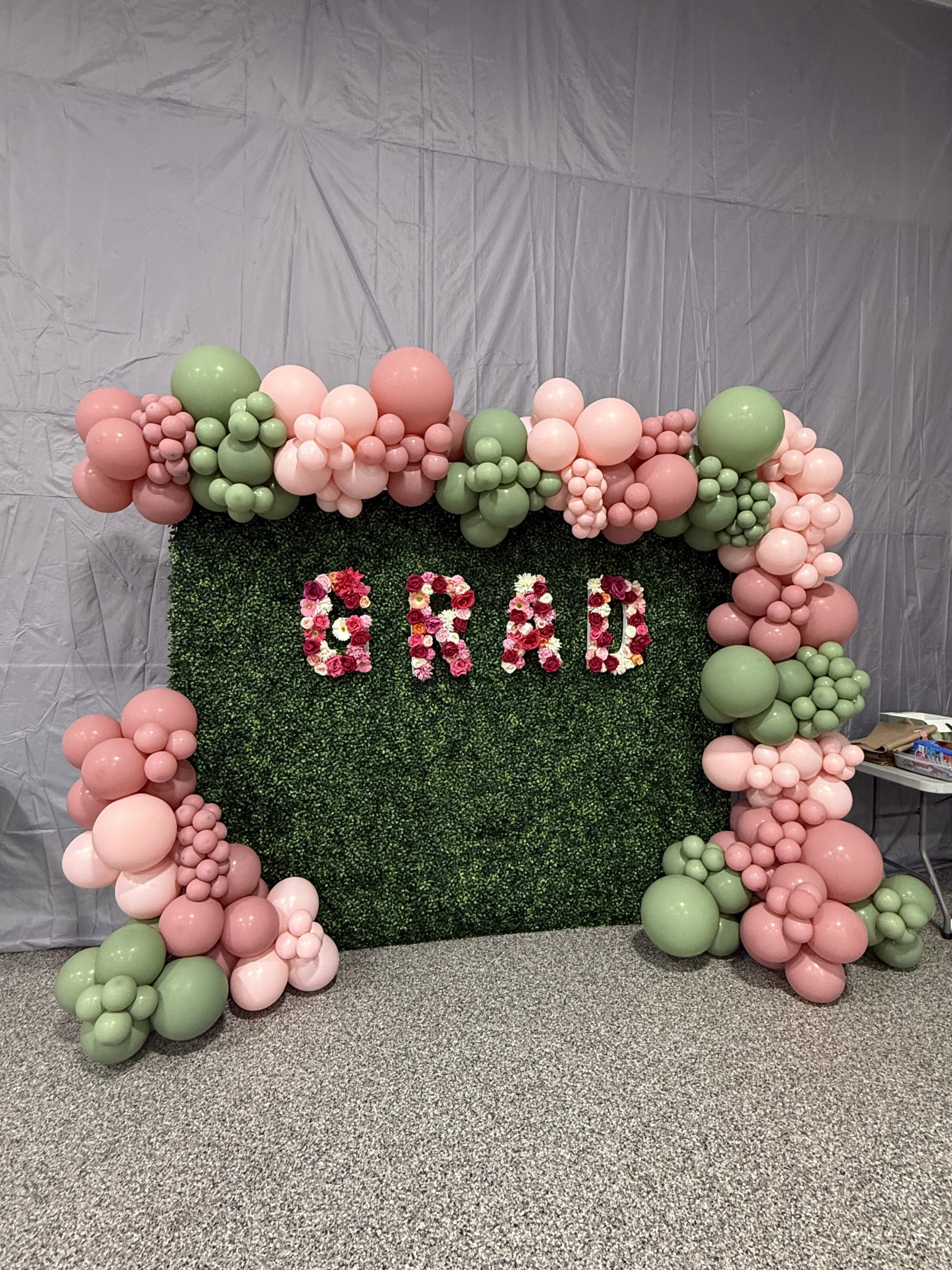 Decorative balloon arrangement with pink and green balloons surrounding a green hedge backdrop with the word "GIRL" spelled out in pink and white flowers.
