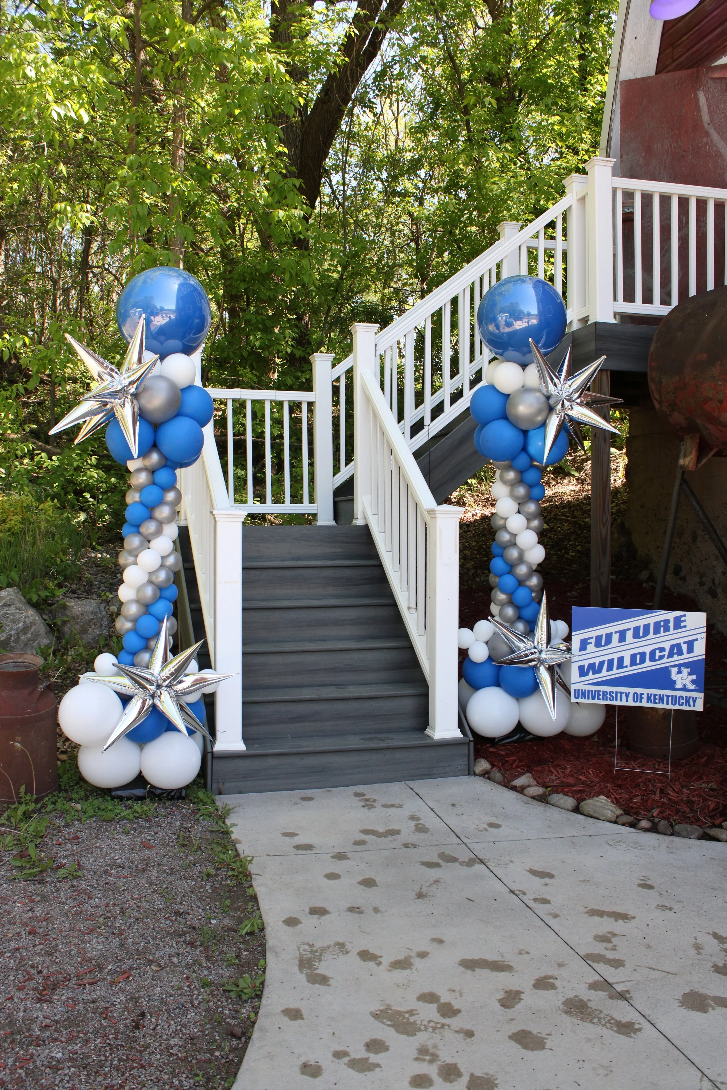 Decorated outdoor stairway with blue, silver, and white star-shaped balloons, and a sign referencing the University of Kentucky Wildcats. Green foliage in the background.