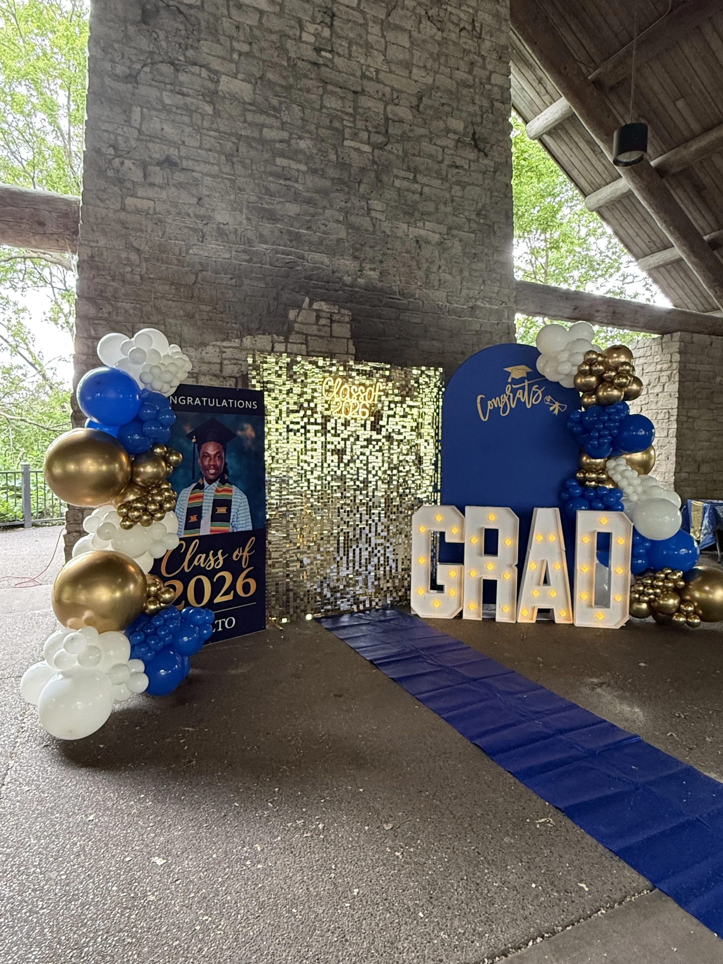 Decorative graduation celebration setup with balloons, a blue walk, and signs reading "Class of 2026" and "Congrats" inside a rustic stone and wood structure.
