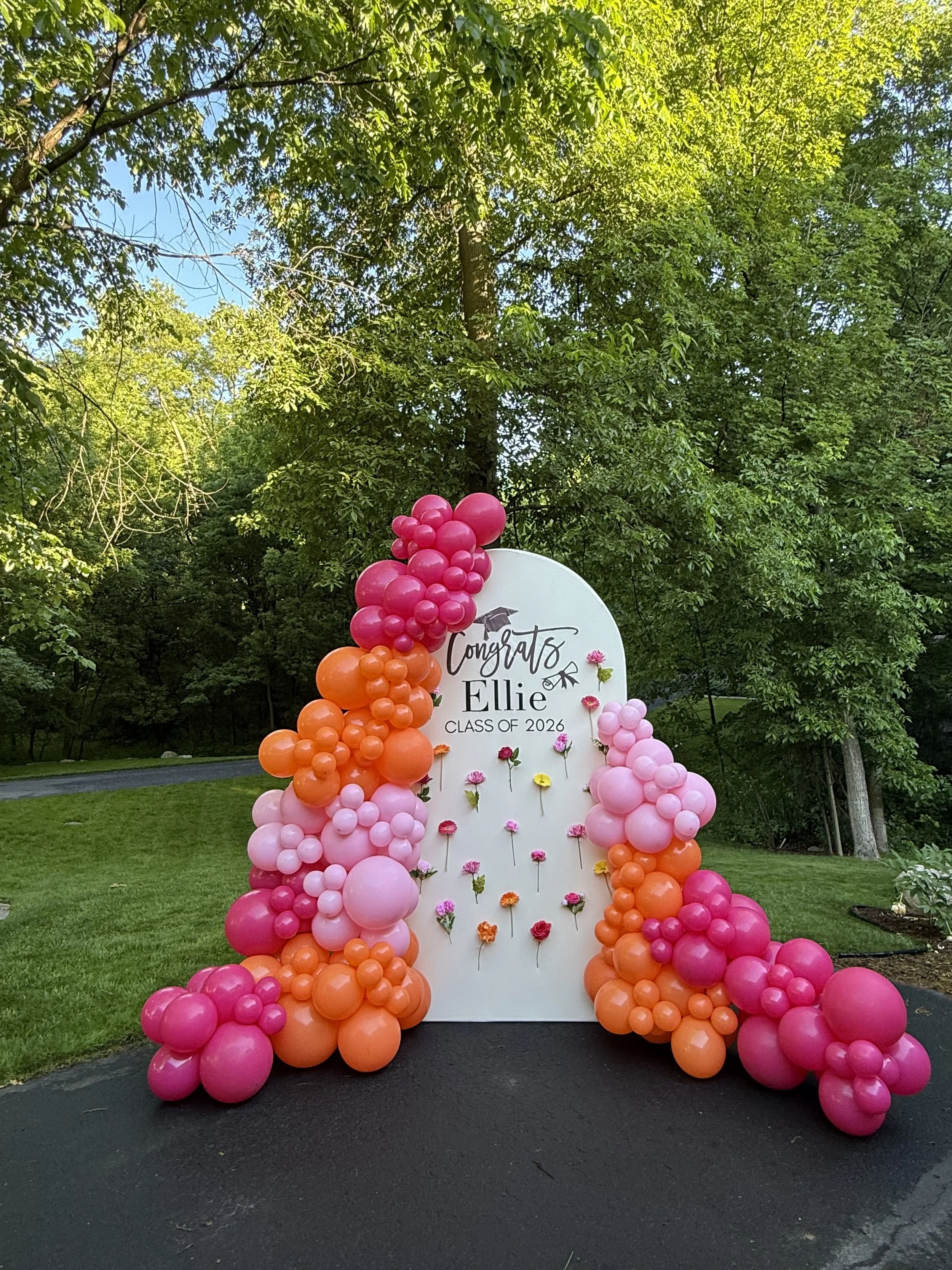 Graduation yard sign surrounded by pink and orange balloons, with flowers attached, on a driveway in front of trees.